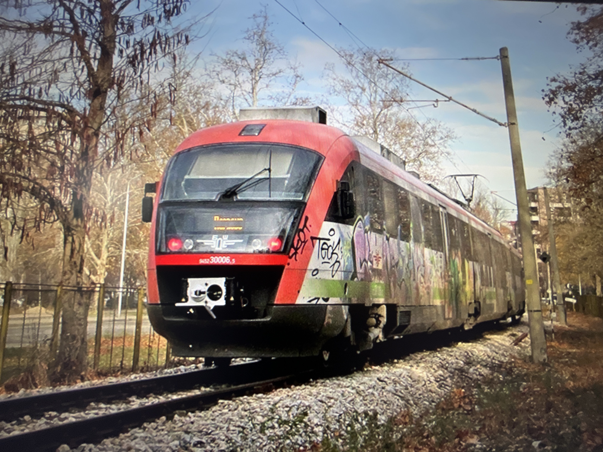 A red and gray electric train with graffiti on its side is stopped on a track in Varna, Bulgaria. The train is in the foreground, with trees and buildings in the background. The sky is blue with some clouds. The train appears to be a passenger train, as it has windows and doors. The graffiti on the side of the train is colorful and varied, with tags and images. The environment appears to be urban, with the train tracks running through a residential or industrial area. The trees are bare, suggesting it is autumn or winter. The overall impression is one of urban decay and neglect, with the graffiti symbolizing an act of vandalism or artistic expression.