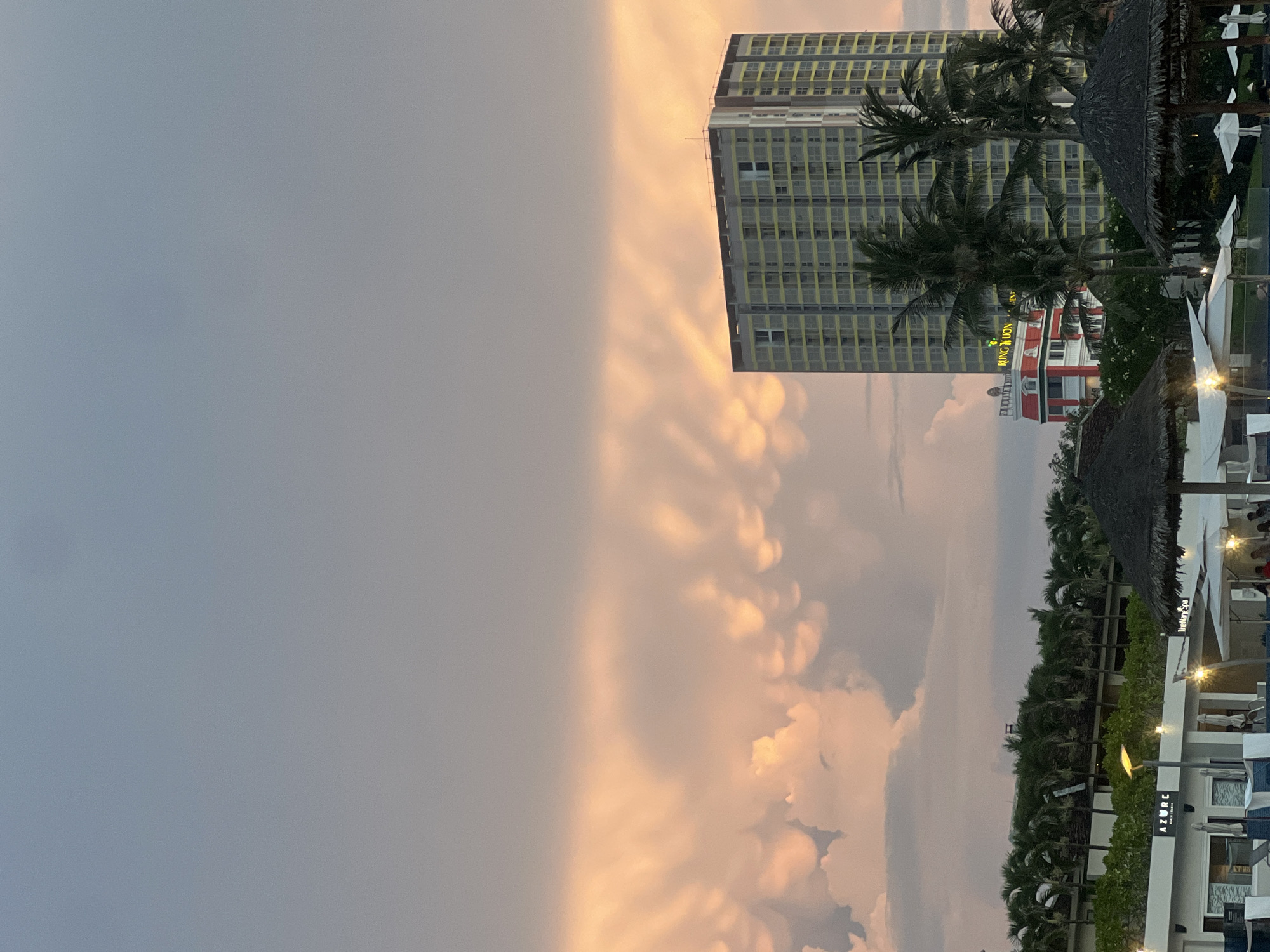 The image captures a tropical resort scene at dusk in Da Nang, Vietnam. The sky is a dramatic canvas of pastel colors, with soft pink and orange hues illuminating the clouds, suggesting either sunset or sunrise. Dominating the background is a tall, modern hotel building with a yellow grid pattern on its facade. In the foreground, lush palm trees sway gently, framing the view and adding to the tropical ambiance.
The scene appears to be a resort area, with elements like thatched-roof cabanas and white umbrellas scattered around. A building with red and white accents and illuminated signage is visible amongst the greenery. There are also glimpses of resort structures, including white buildings with balconies and illuminated signs, one of which reads "AZURE." The overall atmosphere is tranquil and picturesque, evoking a sense of relaxation and vacation. The lighting and cloud formations suggest a peaceful transition between day and night. While people are not clearly discernible, the presence of resort amenities implies guests are present, enjoying the evening.