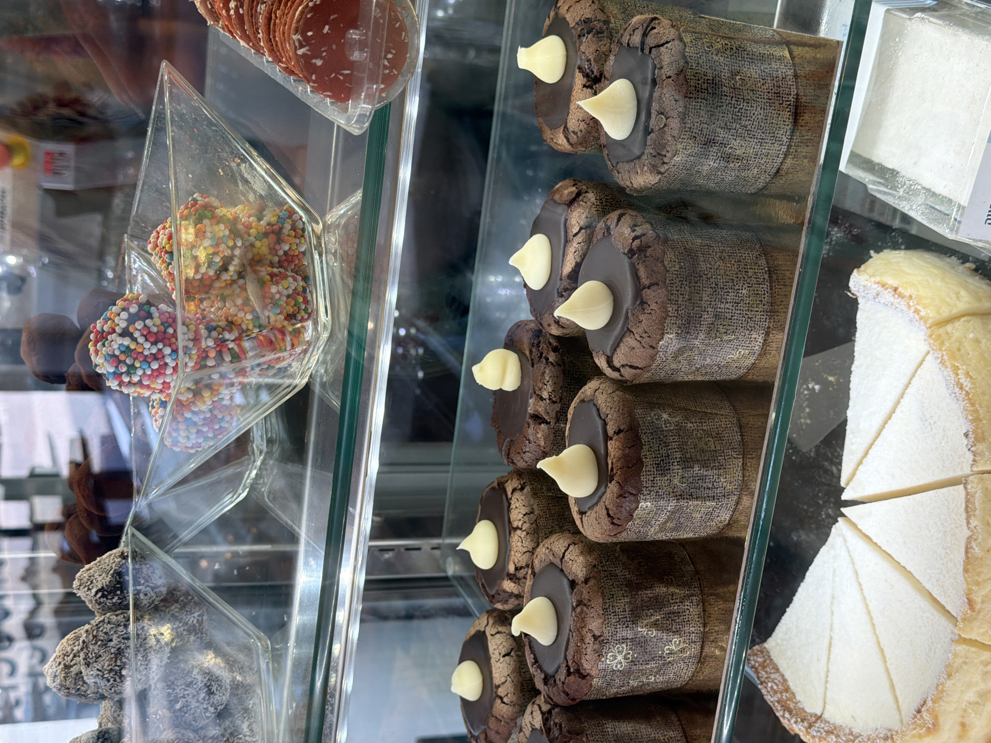 Captured inside a pastry display case in an unknown city, this eye-level shot showcases an array of delectable treats. At the upper left, stacked red wafers sit in a clear container, with rainbow-sprinkled chocolate balls in a triangular case below. Adjacent, there are brown, coconut-covered balls in another triangular case. The right side of the shot highlights rows of chocolate cupcakes with teardrop-shaped white icing in brown-patterned paper containers. Next to them is a sliced, white cake dusted with powdered sugar. The display case is made of clear glass and metal, providing a clear view of the pastries. The lighting is bright, illuminating each item.