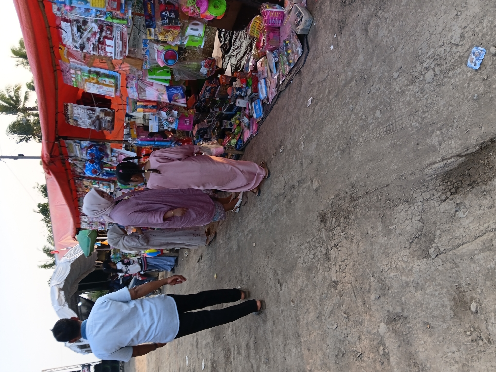 The image depicts a street scene in Sumber Mulyo, Indonesia, possibly a market or roadside stall area. In the foreground, a man in a light blue collared shirt and black pants walks away from the camera, his left hand extended slightly. To his right, a woman in a light purple hijab and long dress walks towards a stall. Next to her, another woman in a darker grey hijab and a long dress is visible. A young girl in a light pink dress, her hair in two braids, stands facing the stall.
The stall itself is covered by an orange tarp and is laden with various items, primarily toys and packaged goods. These are displayed hanging from the tarp and piled on the ground in front of it. Among the items are colorful boxes, plastic packaging, and what appear to be toy vehicles and action figures. The ground in front of the stall is covered with a black tarp. The environment appears to be an unpaved dirt or gravel road, with some tire tracks visible. In the background, a white tent structure is visible, along with palm trees against a clear sky. The overall impression is of an outdoor commercial area.