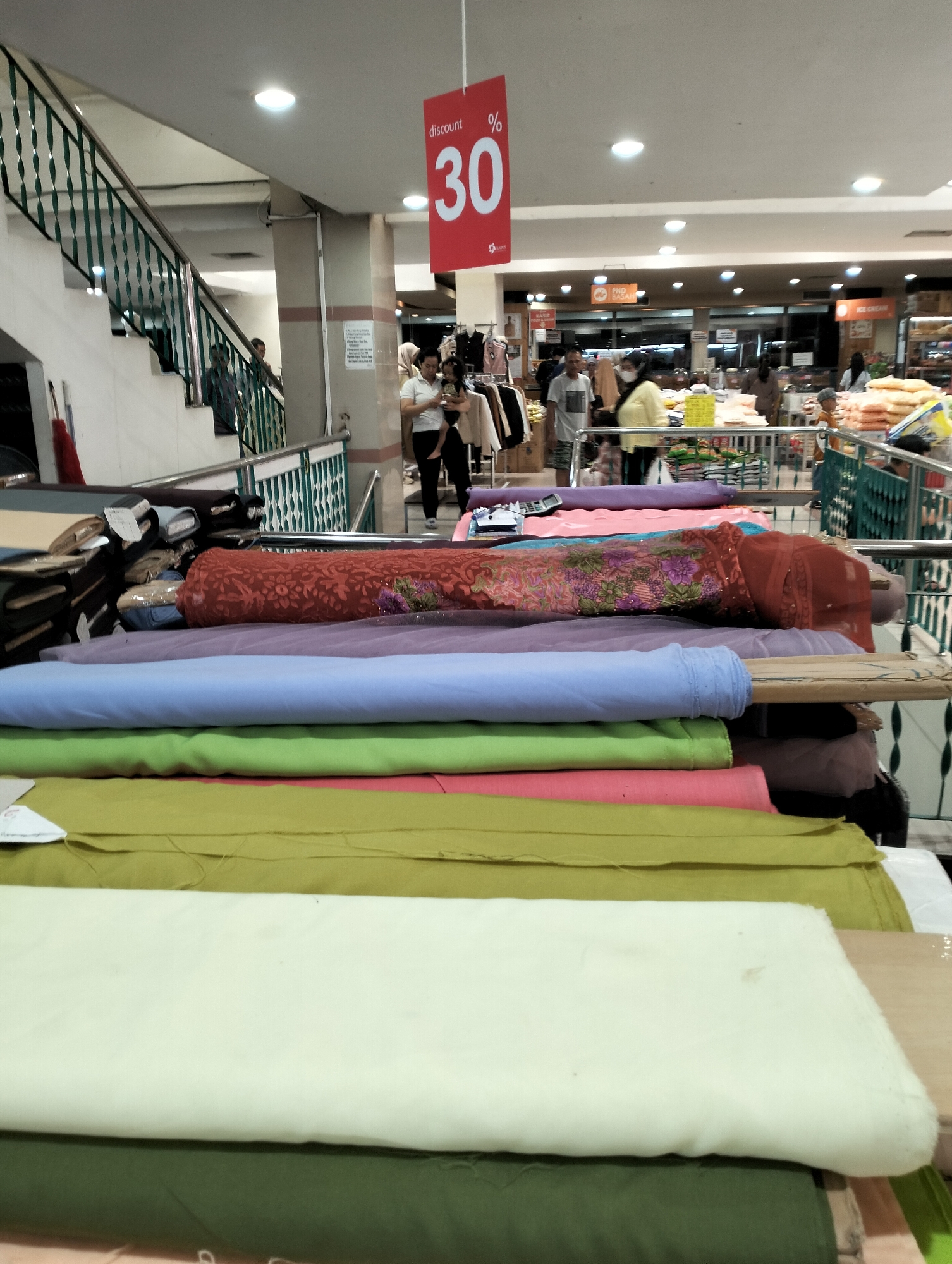 The image depicts the interior of a store, likely a department store or fabric shop, located in Ungaran, Indonesia. In the foreground, a stack of rolled fabrics is prominently displayed, featuring various colors including red, purple, light blue, green, and a light yellow. Behind the fabrics, a staircase with green railings is visible, leading to an upper level. Several individuals are present in the background, some browsing merchandise, others carrying items. A red discount sign with "30%" is hanging from the ceiling. The environment appears to be an indoor retail space with artificial lighting.