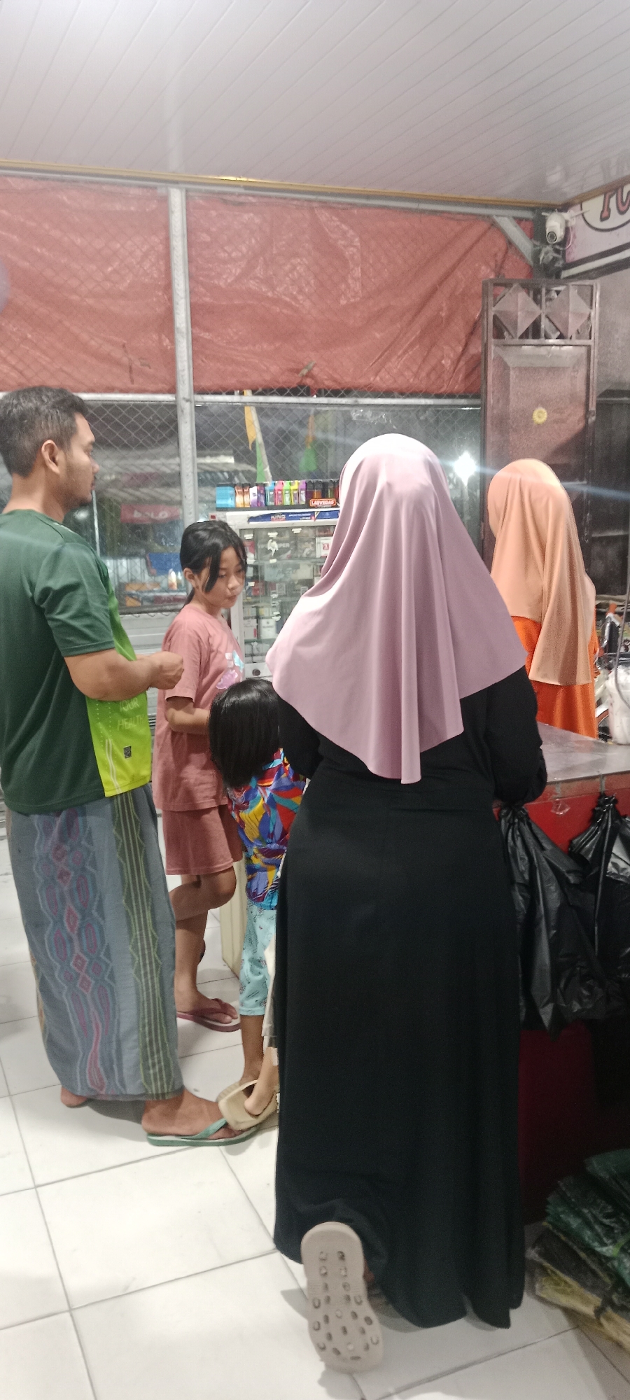 In Rengel, Indonesia, a family is gathered at a convenience store. A man in a green shirt and patterned sarong stands on the left, holding a green bag. To his right, a young girl in a pink shirt looks towards a display of items. Beside her, a child in a colorful shirt is partially visible. A woman in a pink hijab and black dress is in the foreground, facing away from the camera. Another woman in an orange hijab stands further back on the right. The store's interior is visible, with shelves stocked with products and a red counter. The floor is tiled white.