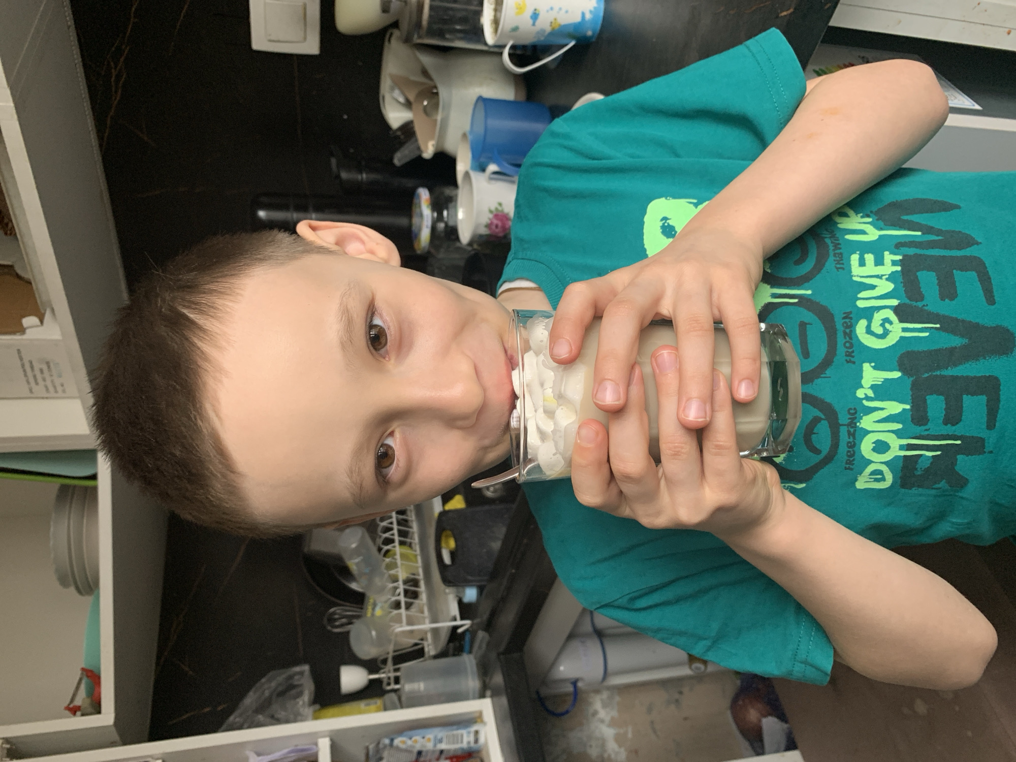 A young boy with short brown hair and light skin is drinking from a glass filled with a creamy liquid and white topping. He wears a green t-shirt with the text "FROZEN DON'T GIVE UP". The background reveals a kitchen environment with a dark countertop littered with mugs and kitchen appliances. A dish rack with bottles sits to the left, and white shelves are visible below. The location is likely a residential kitchen, with no explicit indication of being in Izhevsk, Russia, visible in the image.