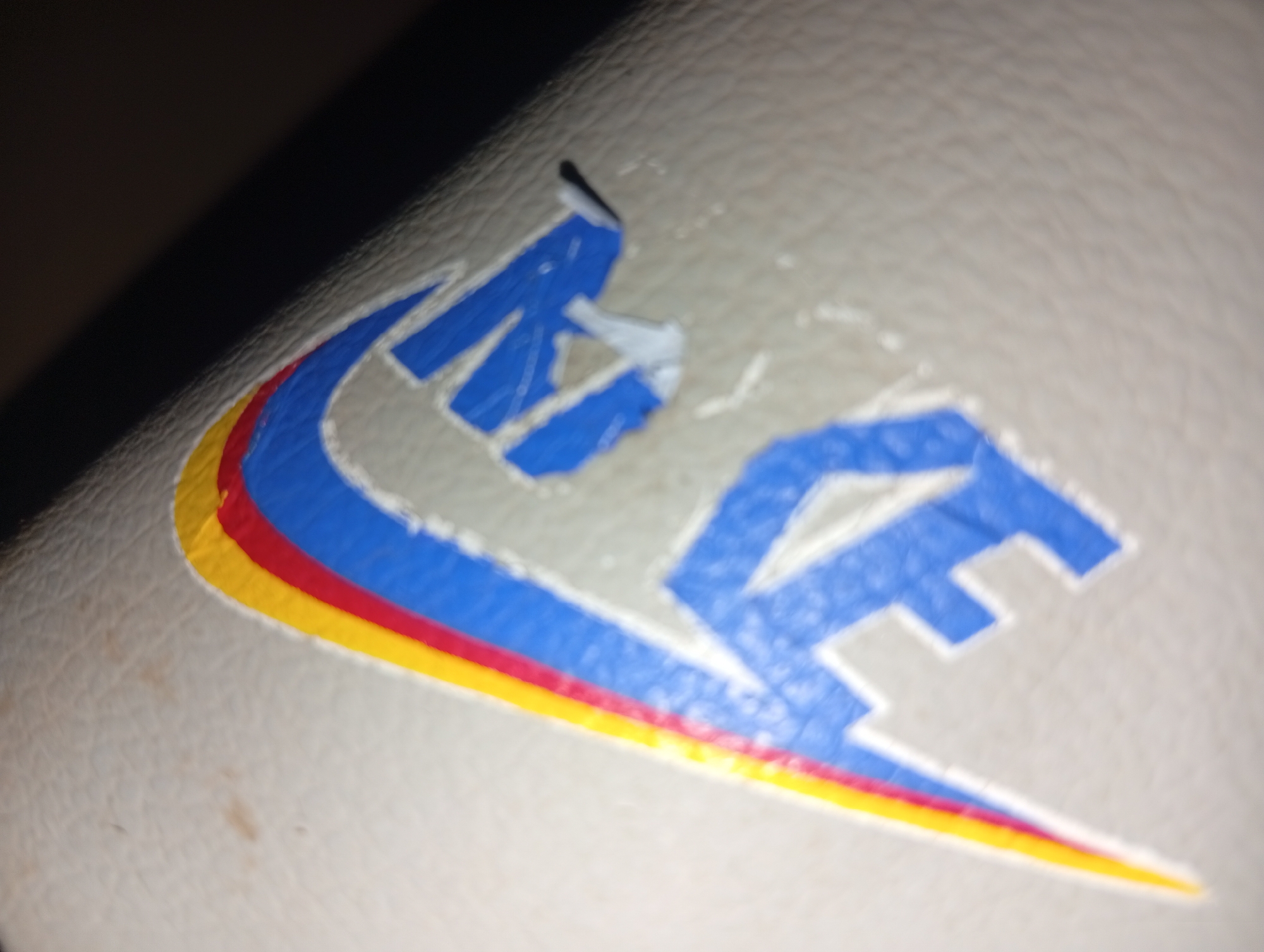 The image shows a close-up of a logo on a textured, light-colored surface, possibly leather or faux leather. The logo is comprised of stylized blue letters, outlined in white. A curved stripe of yellow, red, and blue runs along the bottom left of the logo, suggesting a flag or emblem. The blue lettering appears to be partially worn or damaged, with some areas peeling away. The background is dark and out of focus, suggesting the item is illuminated by a single light source. There is no indication of the city of Ukukwa, Nigeria, in the image.