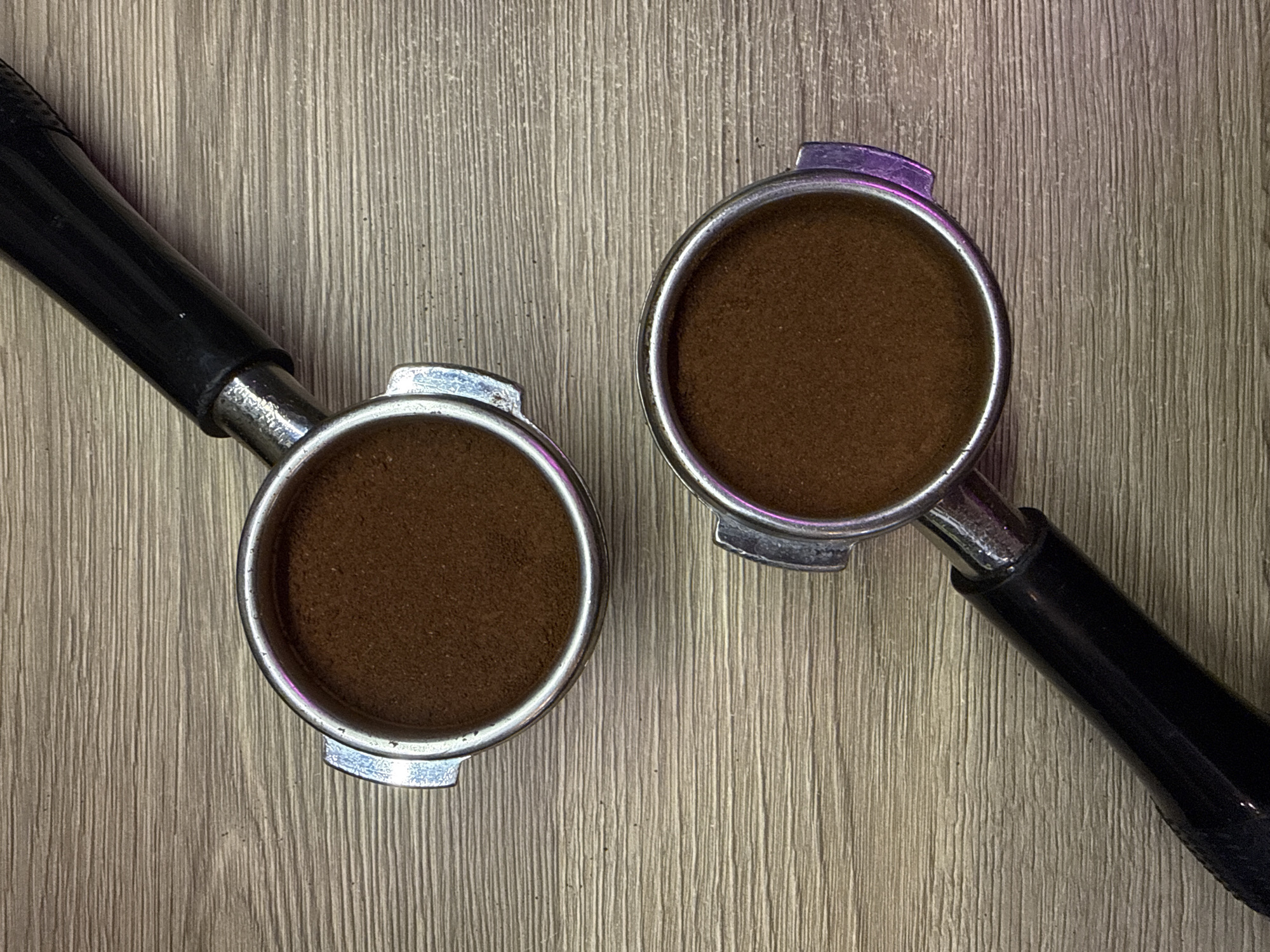 The image shows two espresso machine portafilters filled with ground coffee, resting on a light brown, vertically lined surface, potentially wood or laminate. The portafilters have black handles and silver-colored filter baskets. No information about the location in the city of Khimki, Russia, can be inferred from the image.