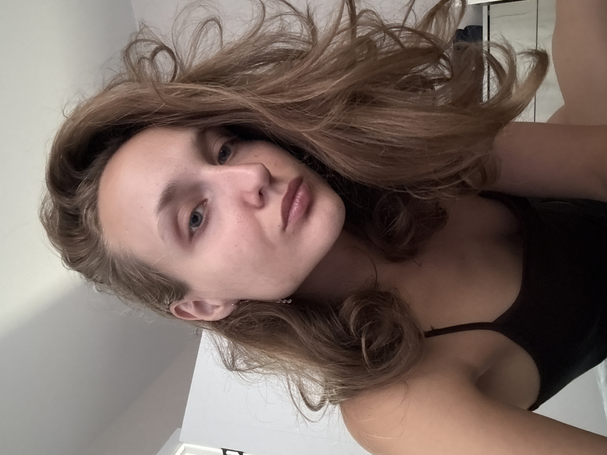 This is a close-up, low-angle selfie of a young woman with light brown hair, taken indoors. She is wearing a dark-colored camisole. Her hair is styled loosely and falls around her face and shoulders. Her facial expression is neutral. The background appears to be a room with a white wall and ceiling, and a glimpse of white furniture. The image was taken in the city of Podshivalovo, Russia.