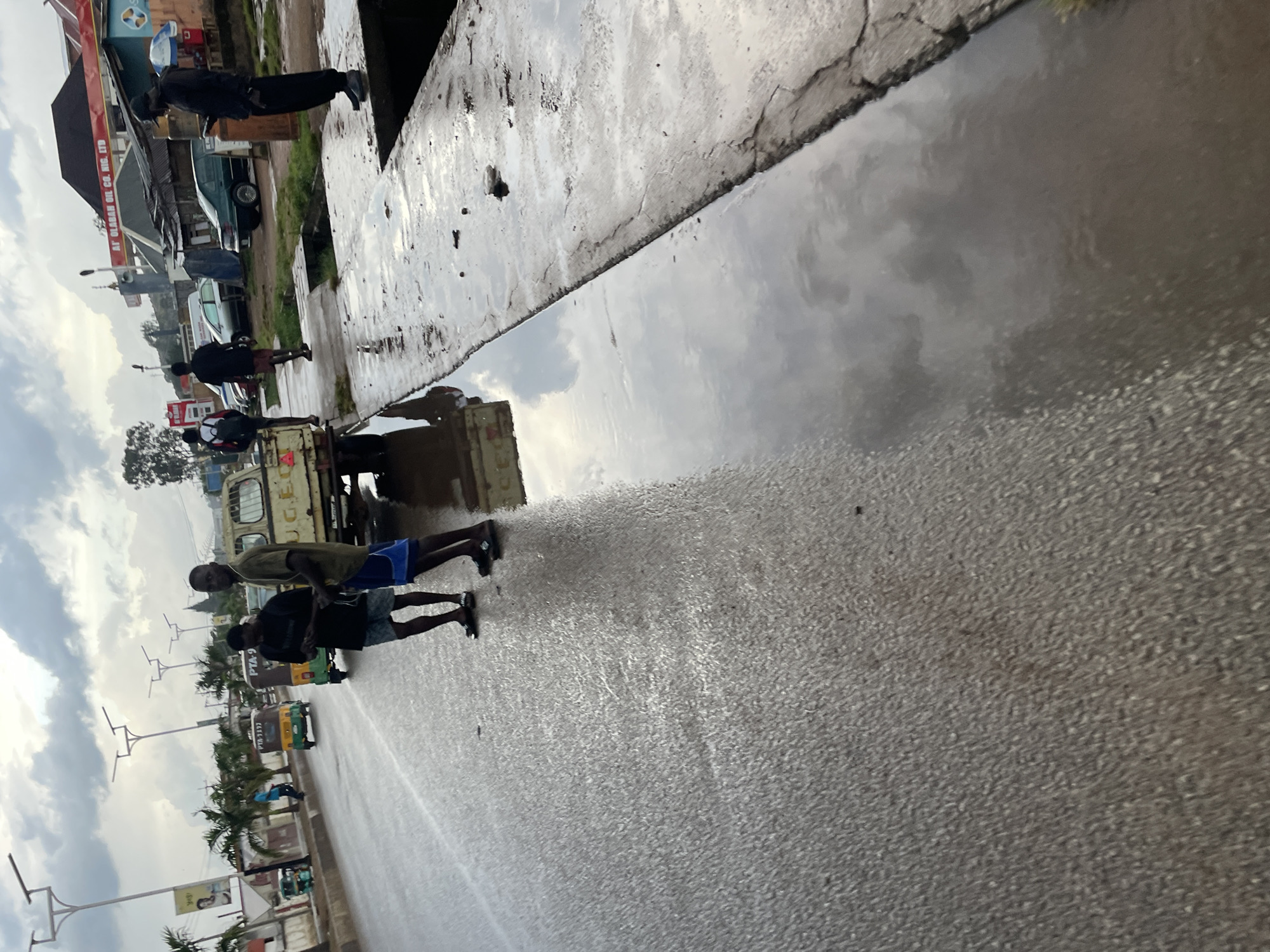 A wide shot shows a street scene in Barakin Lerawi, Nigeria, after a rainfall. The asphalt road is wet and reflects the cloudy sky. Several people are present. In the foreground, two young men stand on the side of the road. One is wearing a black t-shirt and shorts, and the other is wearing a green t-shirt and blue shorts. Behind them, a yellow pickup truck with the letters "U.G.E." visible on its side is parked. Further down the road, a yellow and green motorized tricycle is in motion, and another similar vehicle is parked. In the background, palm trees line the street, and buildings, including a gas station with signage for "AT GLORAH OIL CO. NIG. LTD," are visible. A person in dark clothing is standing on a raised concrete platform, looking at their phone. The overall impression is one of a typical urban street scene in Nigeria, with signs of recent rain.