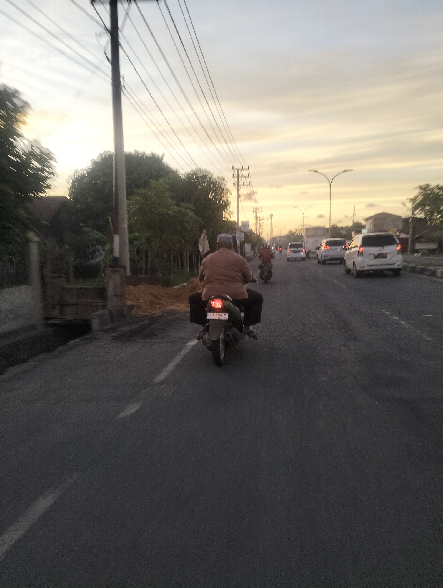 The image depicts a street scene in Lhokseumawe, Indonesia, during the golden hour. The sky is a soft gradient of yellow and white, indicating either sunrise or sunset. In the foreground, a person is riding a motorcycle. The rider is wearing a brown shirt and dark pants, and a white cap. The motorcycle has a license plate that appears to read "BL 5786 NT." The road is asphalt and has a white line marking a lane.
Further down the road, other vehicles are visible, including several cars and at least one other motorcycle. The cars are predominantly white, with one appearing to be a minivan or SUV. The scene is lined with trees and vegetation on the left side, and buildings and streetlights on the right. A utility pole with multiple wires stands prominently on the left side of the road, close to the foreground. There is also a construction area on the left, with a pile of dirt. The overall mood is calm, with the soft light of the sky contributing to a peaceful atmosphere. The motion blur suggests the photo was taken from a moving vehicle.