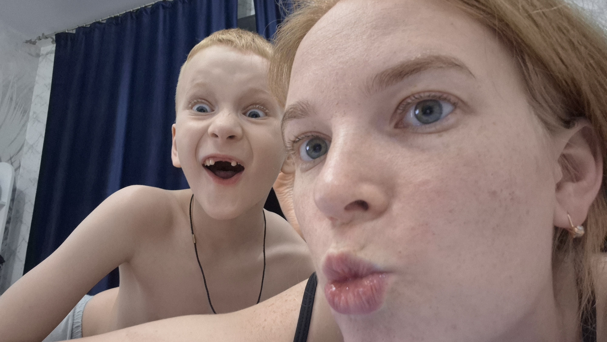 This is a close-up, wide-angle selfie featuring an adult female and a young boy, both making playful expressions. The shot is taken indoors, likely in a casual home setting.
In the foreground, slightly to the right, is the adult female, appearing to be taking the photo. She has reddish-blonde hair, fair skin with visible freckles across her cheeks and nose, and striking light blue or grey eyes. Her lips are pursed into a "duck face" or kissy face. She is wearing what appears to be a dark strap, possibly from a tank top or swimsuit. A small, simple gold hoop earring is visible in her right earlobe. Her expression is lighthearted and humorous.
Behind and to the left of the female is a young boy, also with reddish-blonde hair and fair skin. His eyes are wide open, gazing intently towards the camera, and his mouth is open in a broad, exaggerated smile or scream, revealing a gap where a front tooth is missing. This gives him a very comical and energetic expression. He appears to be shirtless, showing his bare shoulders and chest, and is wearing light grey shorts or underwear. A thin, dark necklace with a small, light-colored bead or pendant is visible around his neck.
The background features dark blue curtains on the right side, suggesting a window or room divider. To the left, there's a lighter wall with a subtle patterned wallpaper, possibly white with faint grey designs. A white object, possibly a piece of furniture or an appliance, is partially visible on the far left. The lighting is soft and even, typical of indoor artificial light or diffuse natural light. The overall mood of the image is joyful, spontaneous, and playful, capturing a candid moment of fun between the two individuals.
