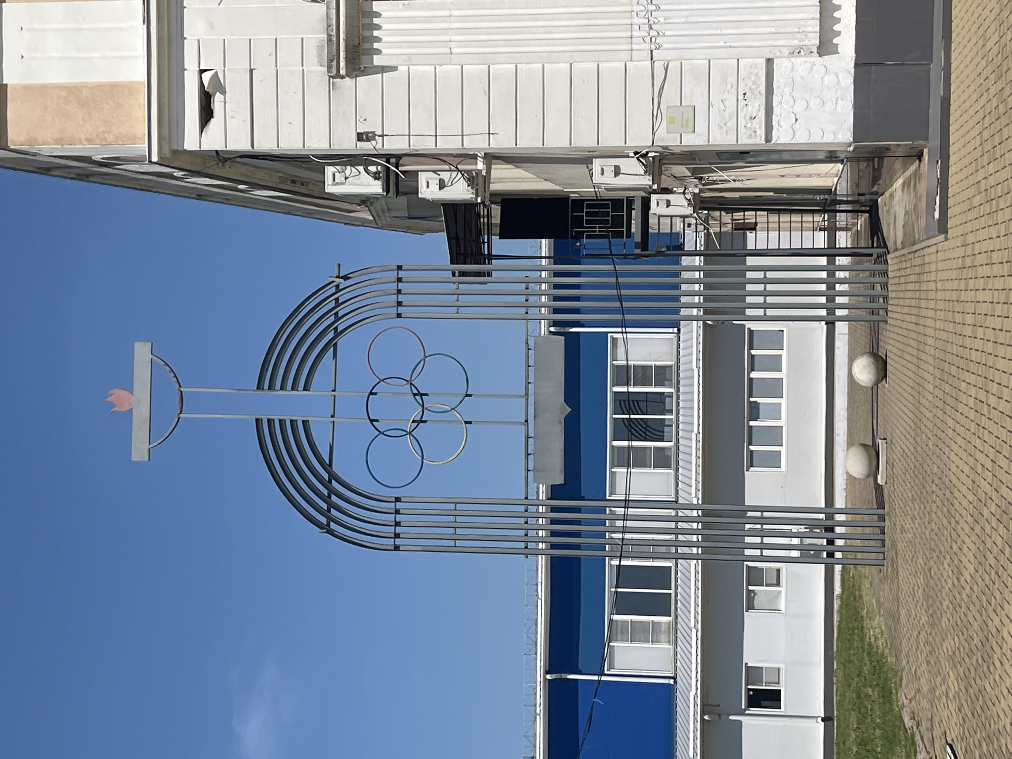 This image, taken from a low angle, shows a metal sculpture resembling Olympic rings and a torch against a bright blue, clear sky. The sculpture is situated in front of a modern building with blue and white sections and numerous windows. The building has a distinct, somewhat industrial aesthetic with exposed piping and outdoor electrical units visible. To the right of the building, a light-brown brick pavement is visible, with a strip of green grass between the pavement and the building. Two spherical bollards are placed along the pavement.
The scene appears to be outdoors during the daytime, with strong sunlight casting shadows. The weather is fair and clear. The overall impression is one of a public space, possibly an athletic facility or a community center, given the Olympic-inspired sculpture. There are no people visible in the image. No text is legible in the image. The location context provided is Takhtamukai, Russia.