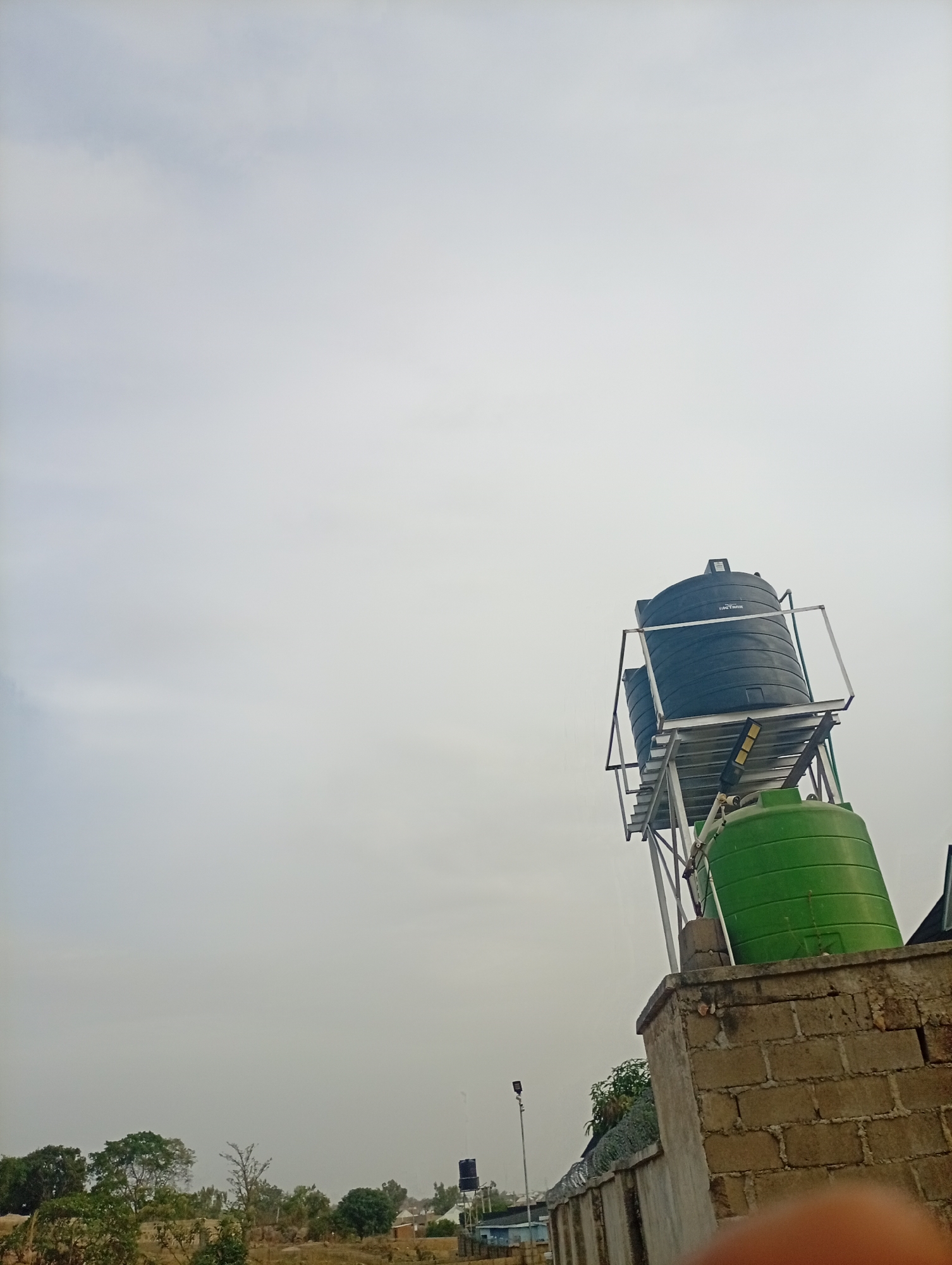 The image shows a water tower in Jos, Nigeria. The tower is constructed of a green tank at the base, with a gray metal frame supporting two black tanks on top. The tower is built on a low wall made of tan-colored bricks. A fence topped with barbed wire runs along the base of the wall. In the background, there are trees, buildings, and a light pole. The sky is overcast. A finger is visible in the lower right corner of the image.