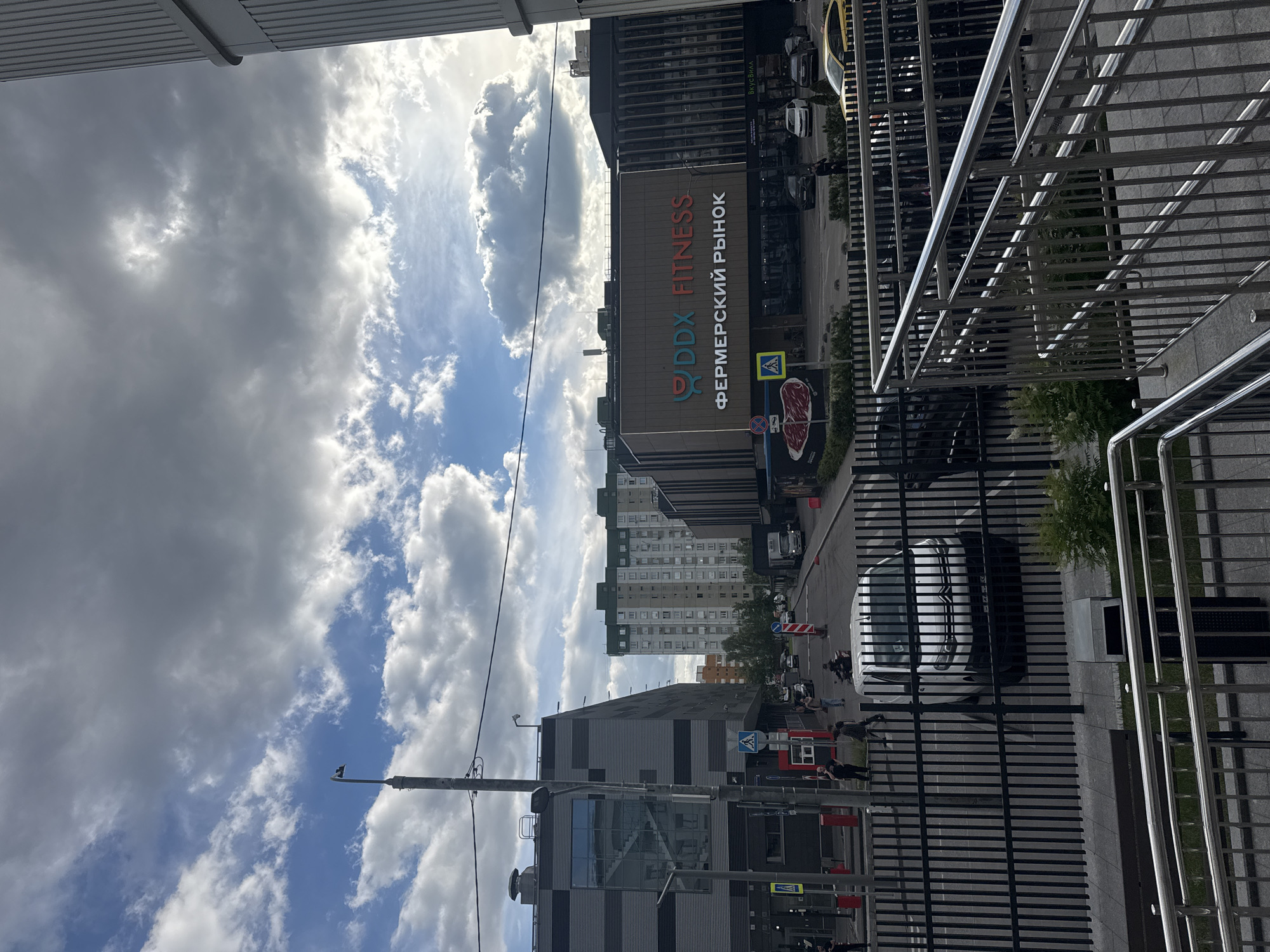 The image shows a street scene in Moscow, Russia. The time of day appears to be afternoon, with bright sunlight filtering through scattered clouds. The sky is a mix of blue and white, indicating partly cloudy weather.
In the foreground, there is a modern building with a metal facade and large windows. A metallic fence runs along the front of the building, separating it from the sidewalk and street. Several cars are parked along the street, and a few people are visible walking or standing.
A prominent feature of the scene is a large sign on the building that reads "DDX FITNESS" in English and "ФЕРМЕРСКИЙ РЫНОК" in Russian, which translates to "FARMER'S MARKET". This suggests the building houses both a fitness center and a market.
Further down the street, more buildings can be seen, including residential apartments with multiple stories. Traffic signs and construction barriers are also present, indicating ongoing activity or maintenance in the area. The overall impression is one of a busy urban environment with a mix of commercial and residential spaces.
