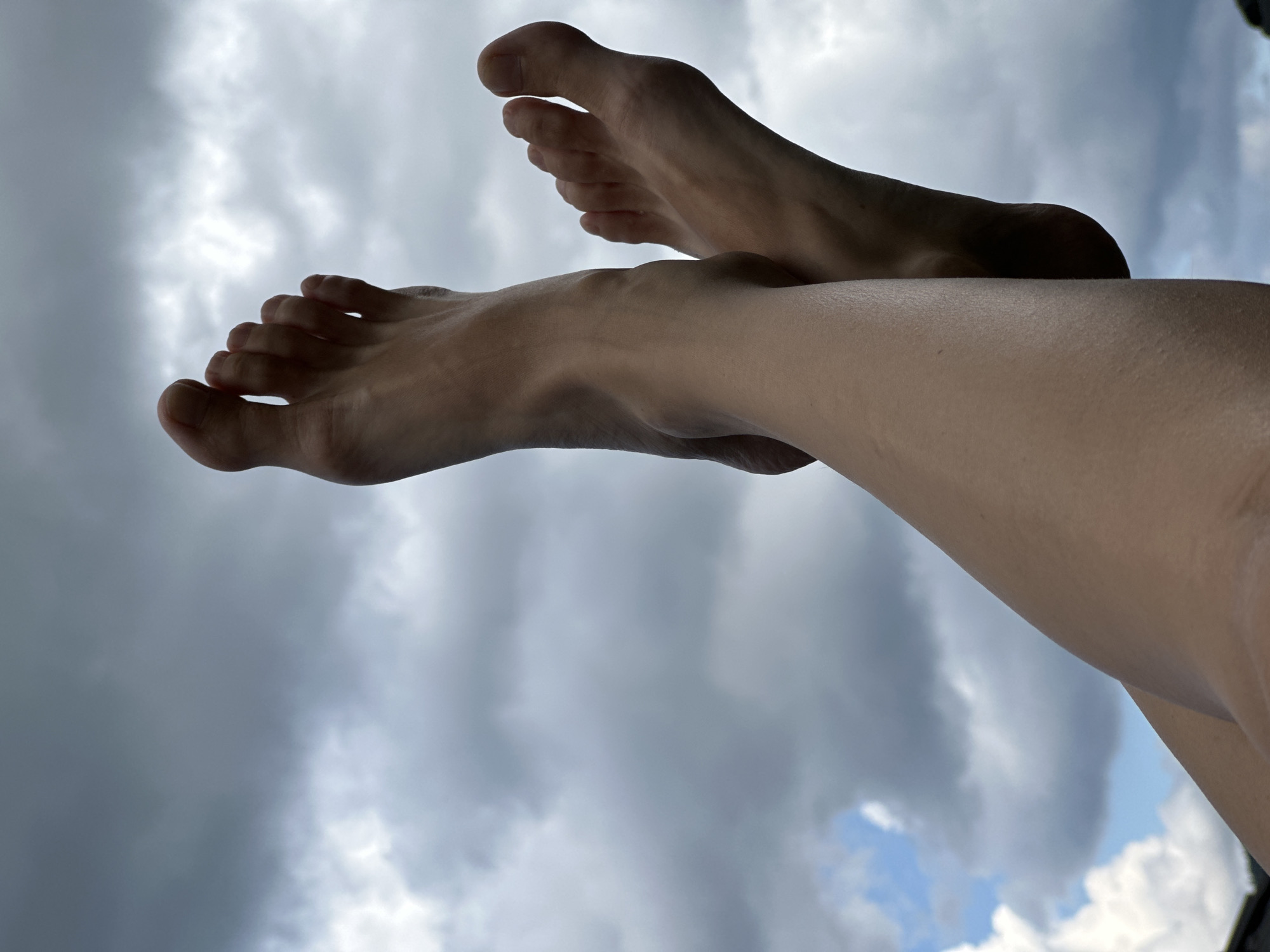 A close-up, low-angle shot captures two bare human feet and lower legs extending upwards against a cloudy sky. The feet are positioned with the left leg crossed over the right, with toes splayed slightly. The skin appears smooth and fair. The background consists of a dramatic sky filled with dark, heavy clouds interspersed with patches of bright white and a hint of blue sky. The overall impression is one of a person relaxing outdoors, with the sky serving as a backdrop. The location in Wisła, Poland is not visually ascertainable from the provided image.