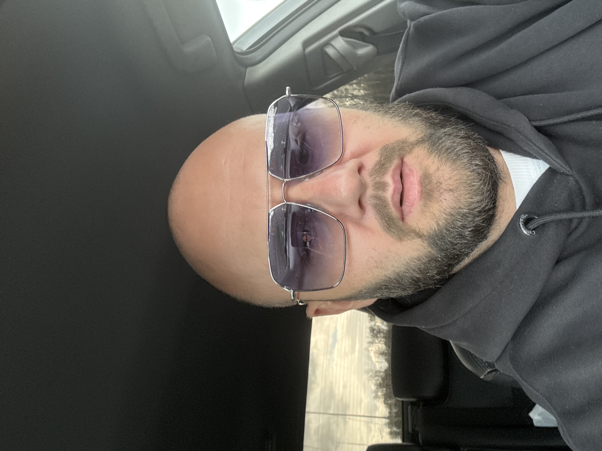 A close-up, slightly rotated image shows a bald man with a beard wearing sunglasses. He is wearing a black hoodie and a white collared shirt is visible underneath. The background suggests he is in a car; the black interior is visible, and the reflection in his sunglasses shows trees and what appears to be a cloudy sky. The location is not discernible from the image.