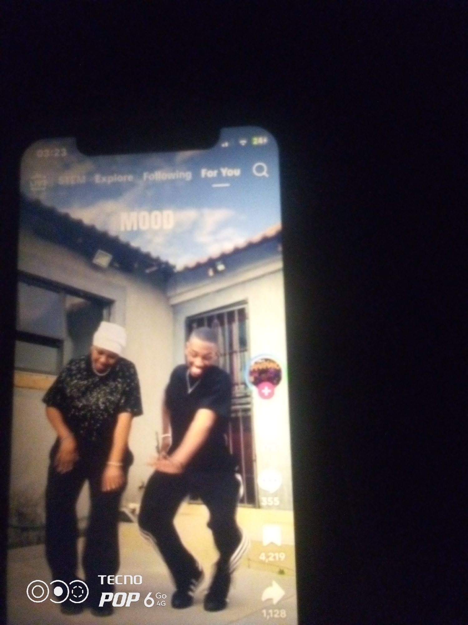 The image shows a smartphone screen displaying a video of two individuals dancing outdoors in front of a building. The building appears to be a single-story house with a light-colored facade and barred windows. The sky above is partly cloudy. On the right side of the screen, there are social media interaction icons and numbers, suggesting a video-sharing platform. In the lower portion of the phone screen, the "TECNO POP 6 4G" branding is visible, indicating the device used to capture or display the image. The specific location within the city of Howayi, Nigeria, cannot be determined from the visual information provided. The individuals are dressed casually, with one wearing a patterned shirt and headscarf, and the other a black t-shirt. Both are in motion, engaged in dancing.