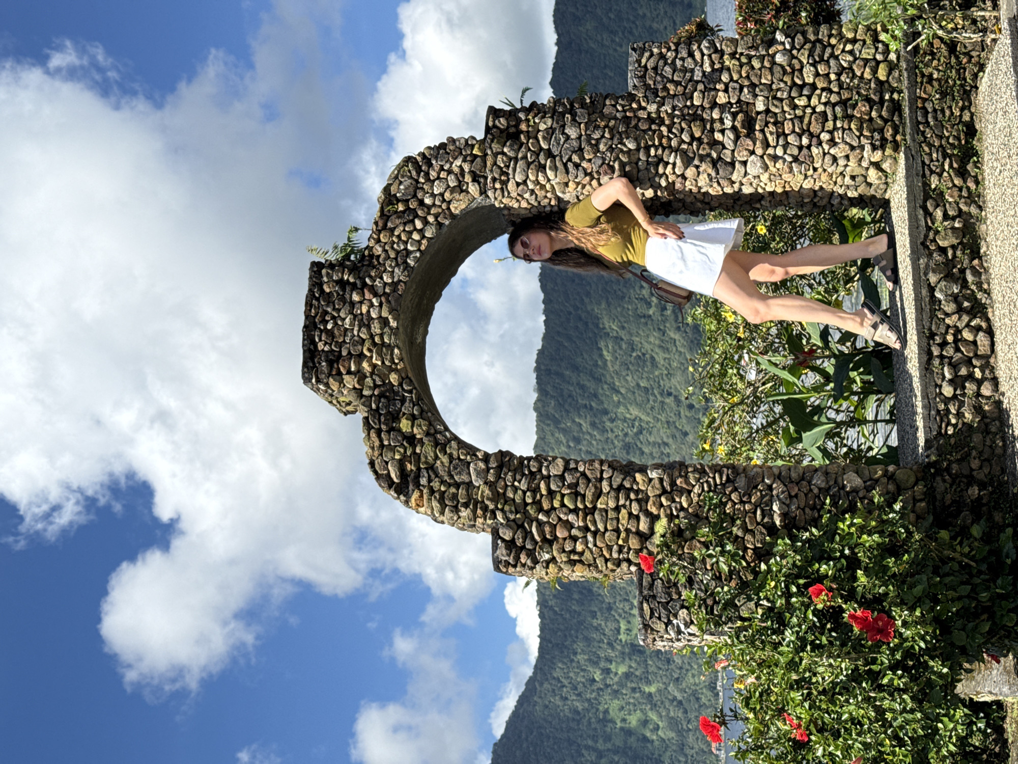 The image shows a woman in Baturiti, Indonesia. She is leaning against a circular stone structure in a garden setting. She wears sunglasses, a green shirt, and a white skirt. A landscape featuring mountains and a blue sky with clouds is visible through the circular stone opening. Hibiscus plants with red flowers are growing along the stone structure. The picture appears to be taken during the day with natural lighting.