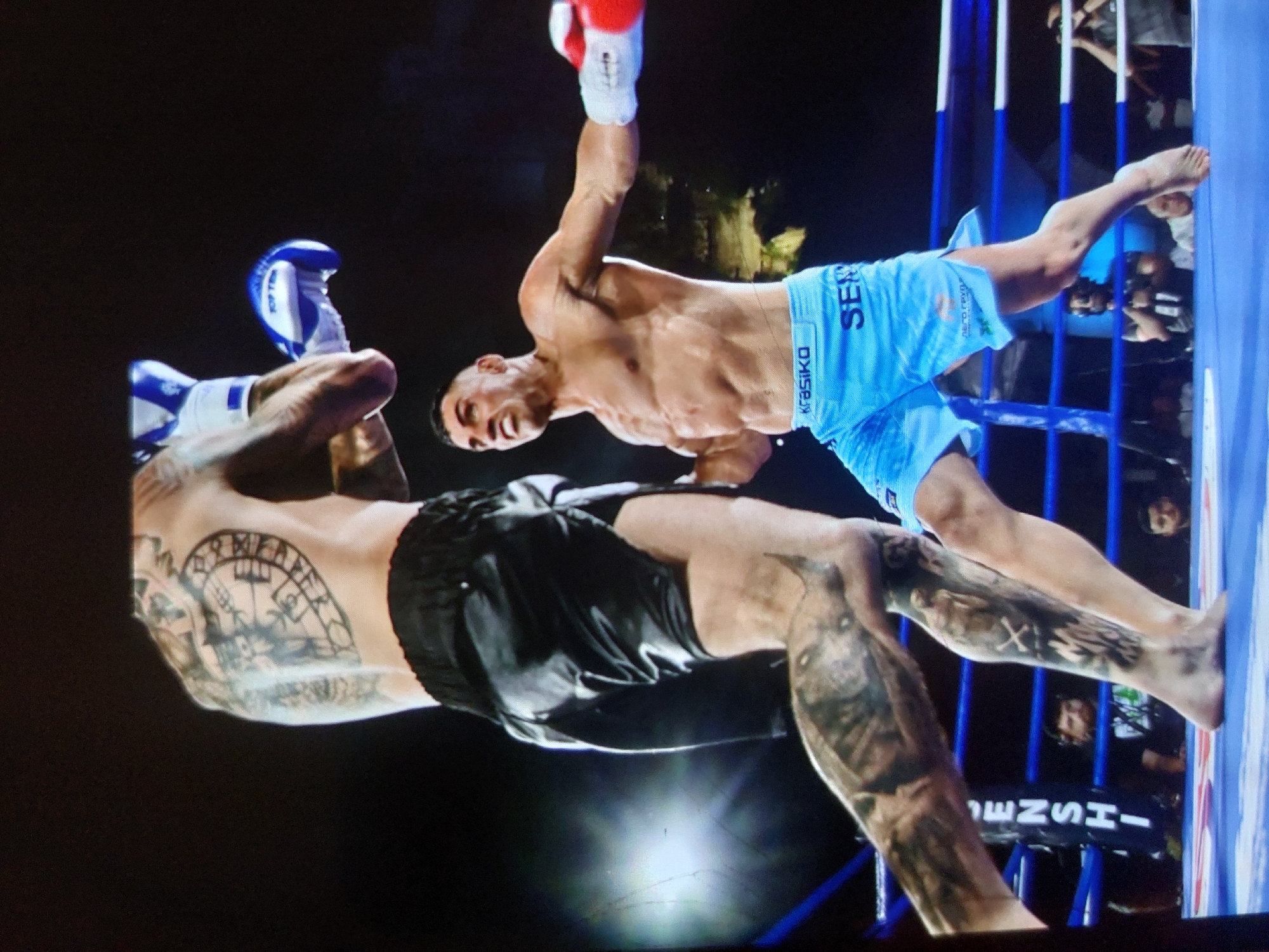 The image captures a dynamic moment during a combat sports match, likely kickboxing or Muay Thai, featuring two male fighters in a ring. The scene is illuminated by artificial lights against a dark background, suggesting a night event. The original image is rotated 90 degrees counter-clockwise.
One fighter, wearing light blue shorts and red and white gloves, is mid-action. He has a muscular, bare torso and is captured with his arm, clad in a red glove, raised high, suggesting a powerful strike or follow-through. His face shows an intense grimace of exertion. Visible text on his light blue shorts includes "SERIK" and "KRASIKO." He is barefoot.
The second fighter, seen mostly from the back, is wearing black shorts and blue and white gloves. He is heavily tattooed on his arms and legs. A prominent tattoo on his upper back/shoulder resembles a Viking compass (vegvísir) or Aegishjalmur, surrounded by what appear to be runes or symbolic patterns. Another tattoo on his leg features an "X" mark. His body is turned, and his arms are raised, indicating he is either reacting to a strike or performing a defensive maneuver. The blue glove has "TOP TEN" visible on it.
The setting is a boxing or kickboxing ring with blue ropes and white posts. Spectators are dimly visible in the background, suggesting a live audience for the event. The intense focus and physical exertion of both athletes are palpable. A banner or board in the background shows partial text, possibly "HENSHE". The event takes place in Tsarevo, Bulgaria.