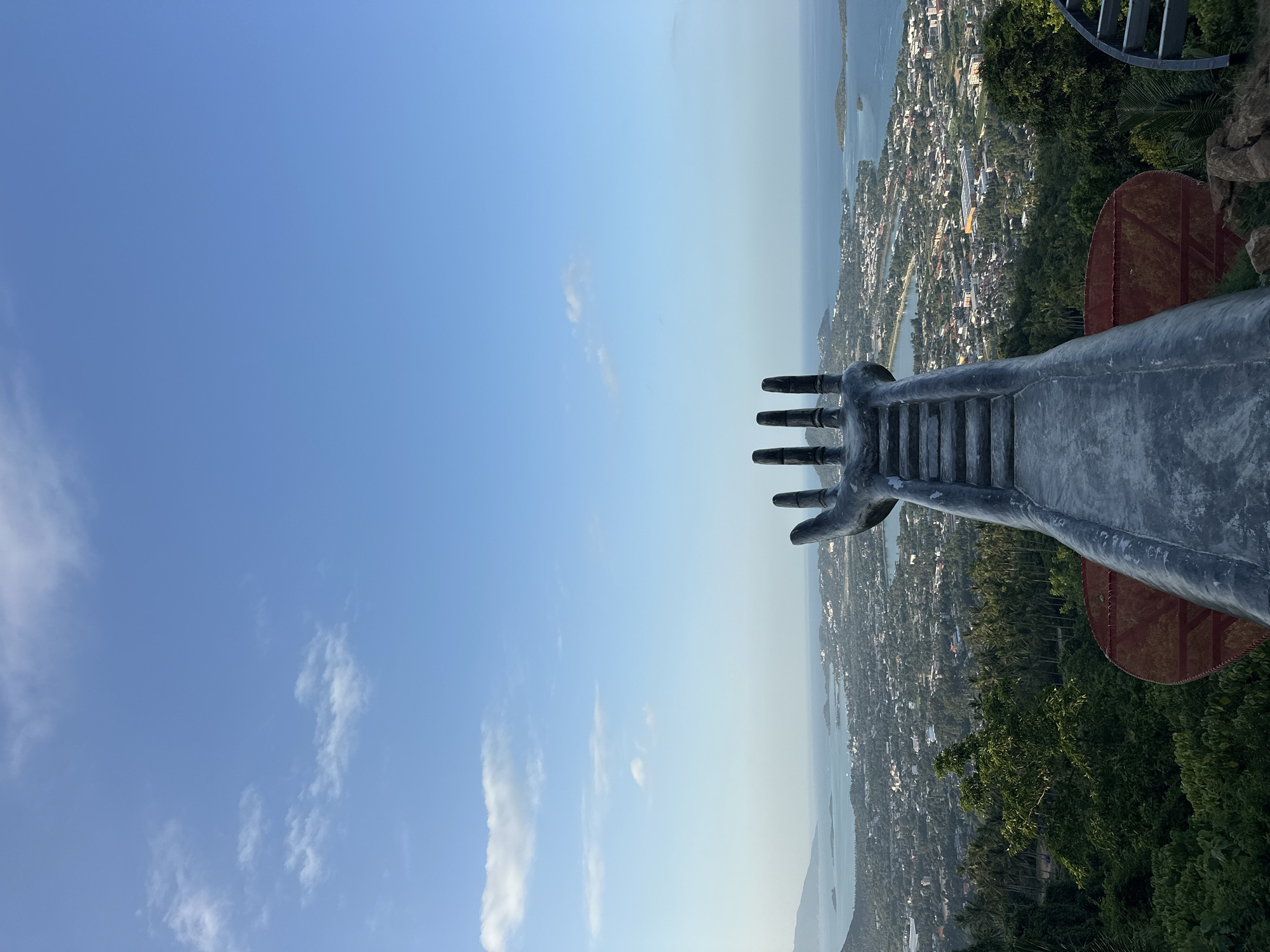 The image, taken from a high vantage point in the Ko Samui District of Thailand, features a monumental statue of a hand reaching towards a sprawling coastal city and the ocean. The statue, made of dark gray concrete, is in the foreground, with its palm facing upwards and its fingers spread, creating a dramatic framing for the landscape beyond. Stairs are visible ascending the forearm of the statue. The city, densely packed with buildings and nestled among lush green hills, stretches out towards the turquoise waters of the sea. Islands dot the horizon, under a clear blue sky with scattered white clouds. The foreground is occupied by the statue and dense green foliage, with some rocks visible at the base. The scene captures a breathtaking panoramic view, emphasizing the scale of both the man-made structure and the natural environment.