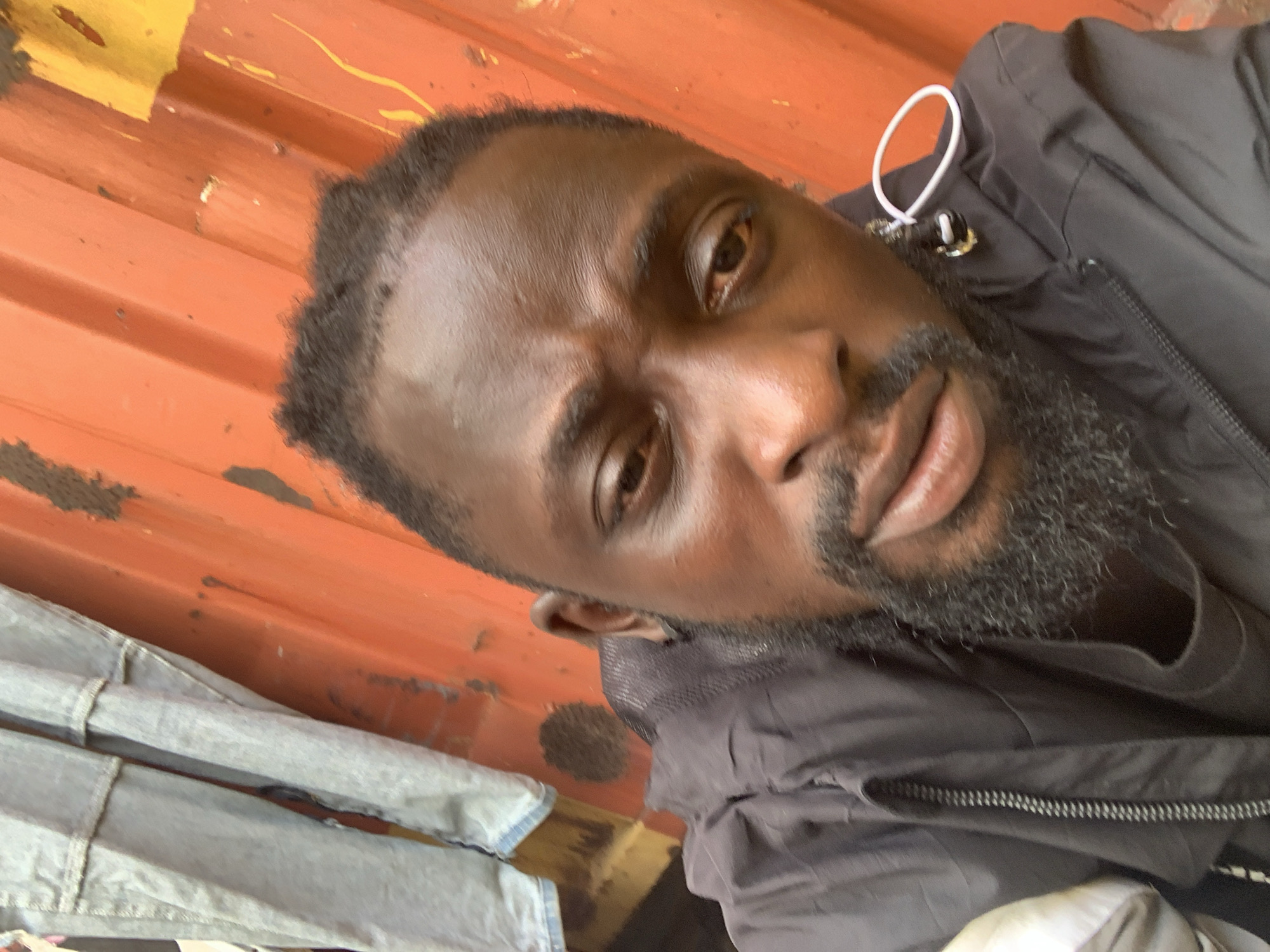 A close-up, slightly tilted portrait features a dark-skinned man in Lekki, Nigeria, looking directly at the camera. He has short, dark hair with a faded style, brown eyes, and a neatly trimmed beard. His expression is neutral to slightly serious.
He is wearing a black jacket with a visible zipper down the front. A white loop, which could be a jacket drawstring or an earbud cord, is visible near his right shoulder.
The background consists of an orange, corrugated metal surface, possibly a shipping container or industrial wall, showing signs of wear and patches of yellow paint. To the lower left of the frame, there is a pile of light-colored fabric, resembling folded denim jeans. The scene appears to be outdoors during the daytime. There is no visible text.