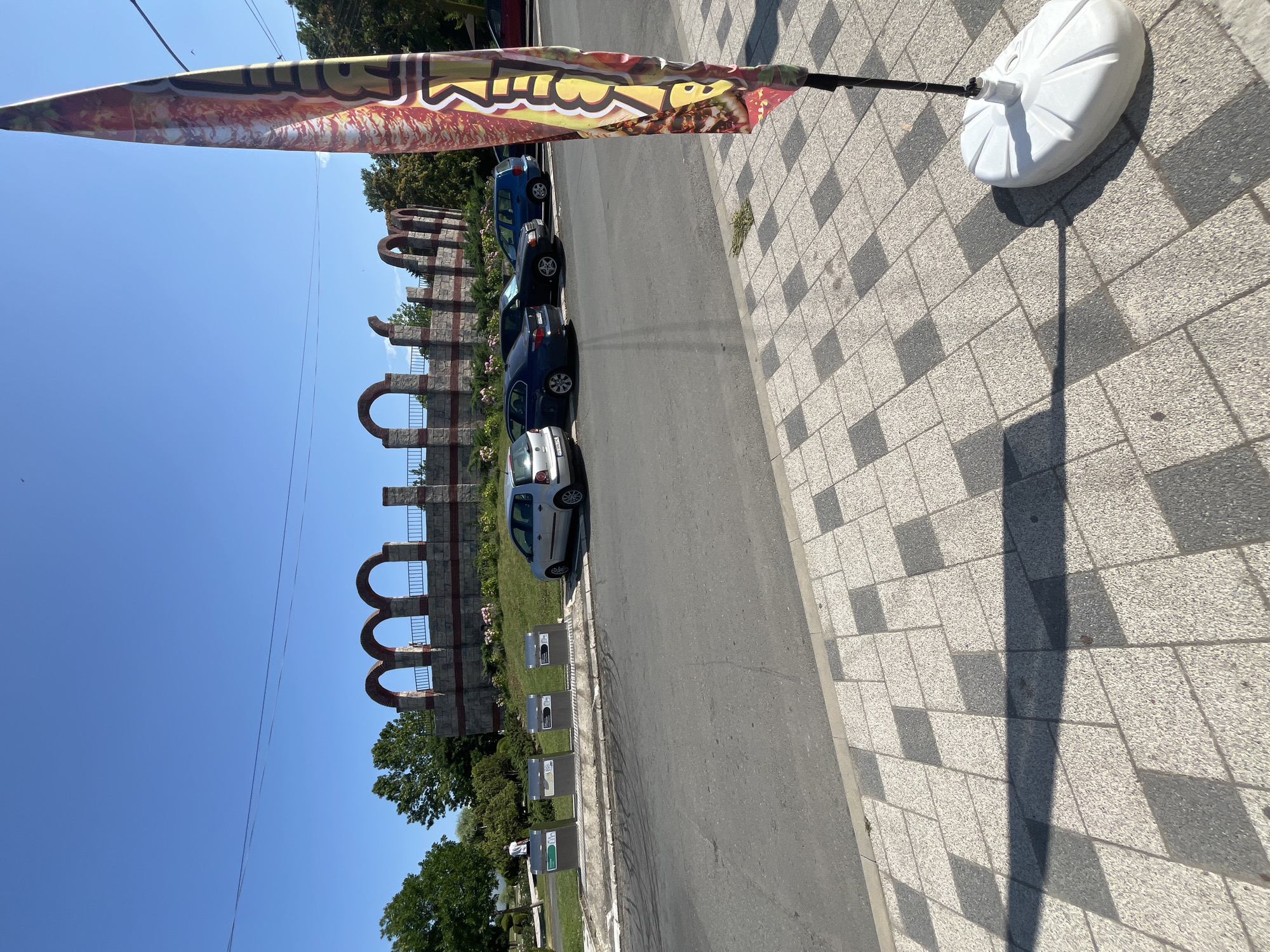 The image shows a street scene in Obzor, Bulgaria, near the Roman Fortress. In the foreground, a sidewalk with a patterned tile design is visible, with a large white base for a flag pole and a colorful flag. Behind the sidewalk is a road with several parked cars, including a silver Volkswagen Polo, a dark blue BMW, and a blue minivan. A section of the road is visible, with tire marks.
In the middle ground, there are several modern, metallic trash receptacles. Beyond the receptacles, a green lawn leads up to a stone structure with multiple arched openings and a decorative railing, which appears to be part of an ancient ruin. A person is visible walking on the lawn in the distance, near the trash receptacles.
The background consists of lush green trees and a clear blue sky. The overall scene depicts a sunny day in a city that blends historical remnants with modern infrastructure.