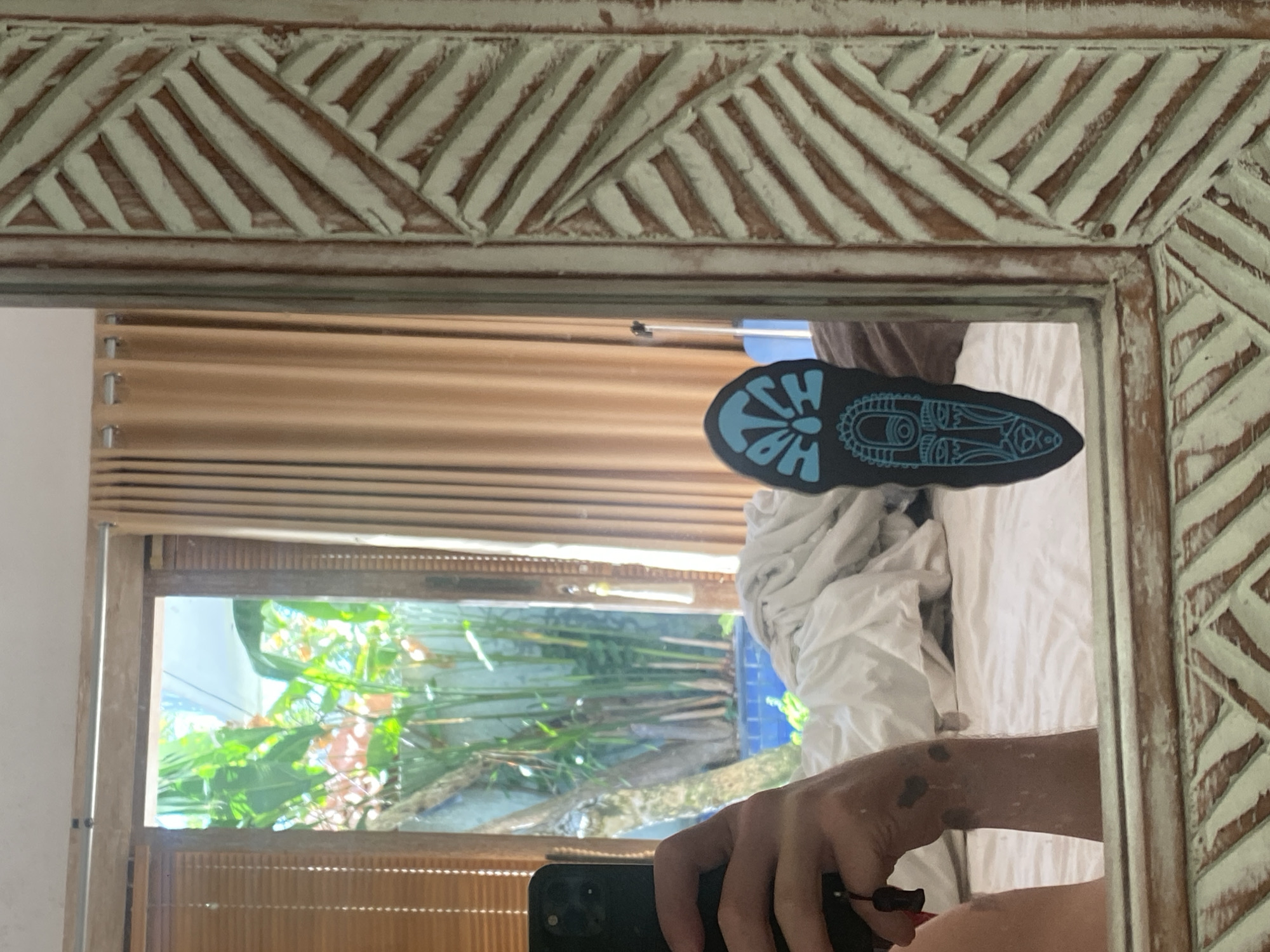 A selfie is taken in a bedroom with a mirror that has an intricately carved wooden frame. The mirror reflects a person's arm and hand holding a black smartphone, and a portion of a bed with white linens. A dark sticker with a tribal mask design in blue is adhered to the mirror. In the reflection, a window shows lush green foliage, suggesting an outdoor tropical environment, possibly a garden or courtyard, with a glimpse of blue water. The background also includes light brown curtains and bamboo blinds. The image was taken in Kuta Selatan, Indonesia.