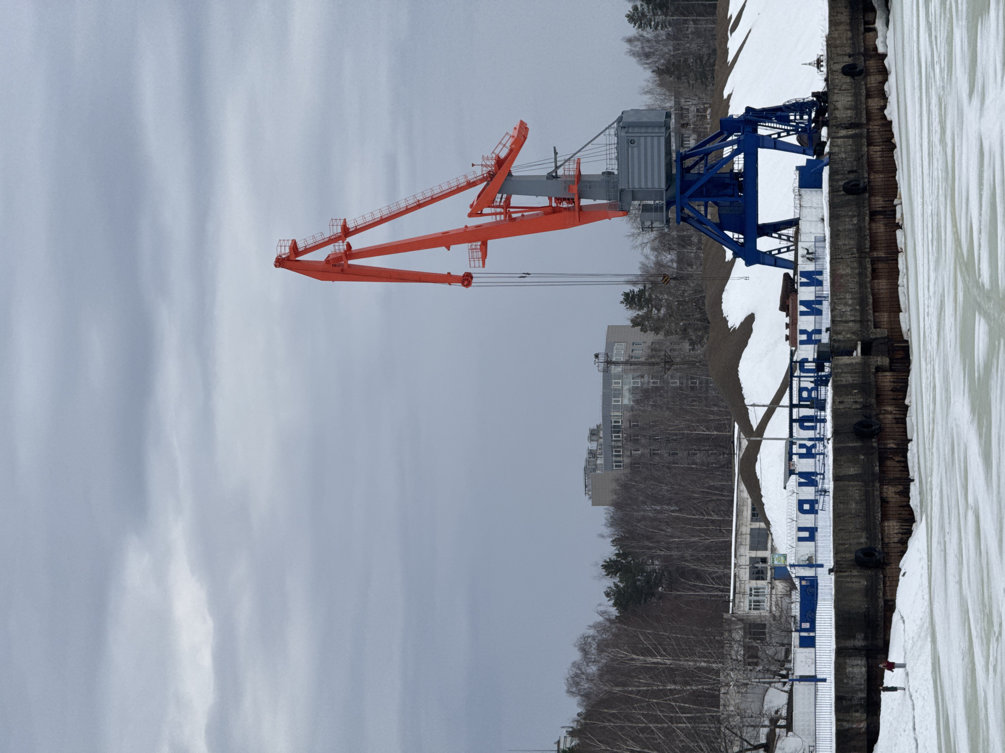 The image depicts a port crane in the city of Chaikovsky, Russia. The crane is orange and blue, and it stands on a concrete dock along a body of water that appears to be partially frozen. The name of the city is written vertically on the dock. Trees and buildings are visible in the background under an overcast sky.