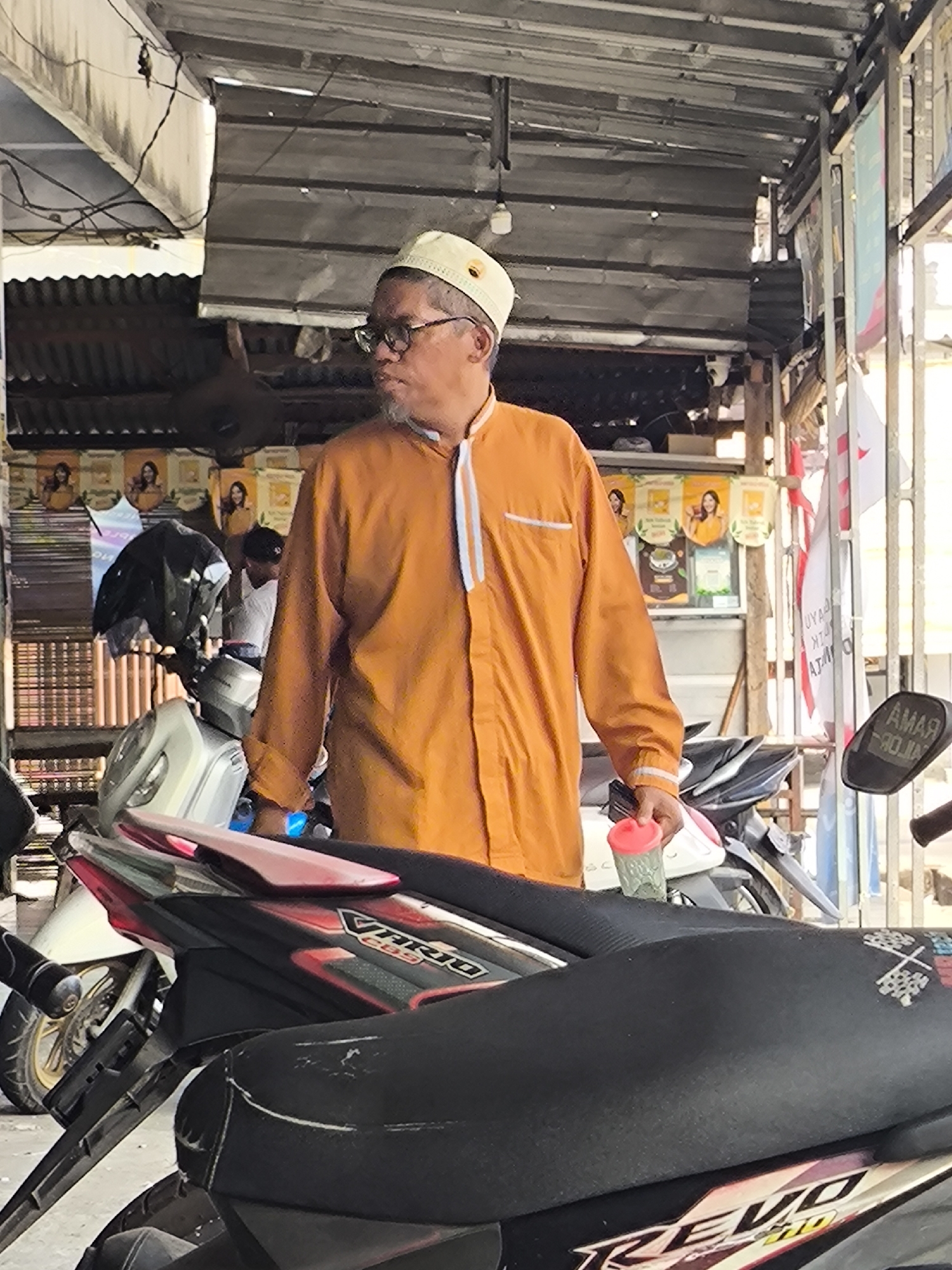 A man wearing a light-colored peci (cap) and glasses stands in front of motorcycles. He is wearing an orange long-sleeved shirt with white trim. The man is holding a pink-capped plastic cup. In the background, there are posters on the wall and various objects stacked. The scene appears to be an outdoor market or shop in Cilegon, Indonesia.