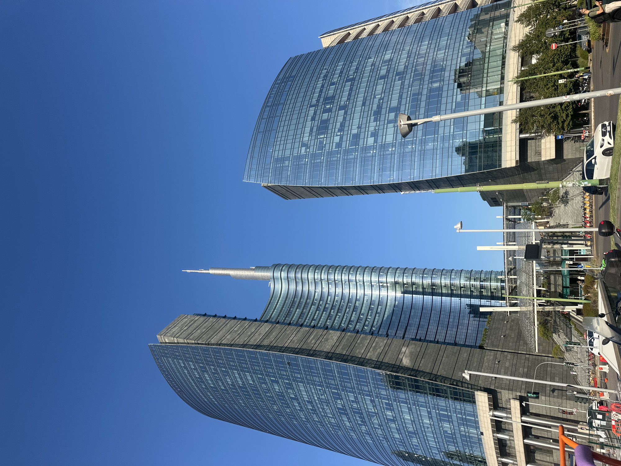 The image shows the modern skyscrapers of the Piazza Gae Aulenti in Milan, Italy, under a clear blue sky. The two prominent buildings are the Unicredit Tower, characterized by its distinctive curved glass facade and spire, and another glass-fronted tower to its right. In the foreground, a street scene unfolds with various vehicles, including a white Mercedes sedan and parked bicycles, suggesting an urban environment. A street lamp with a distinctive modern design is positioned prominently in the middle ground. The overall impression is one of contemporary architecture and a bustling city setting.