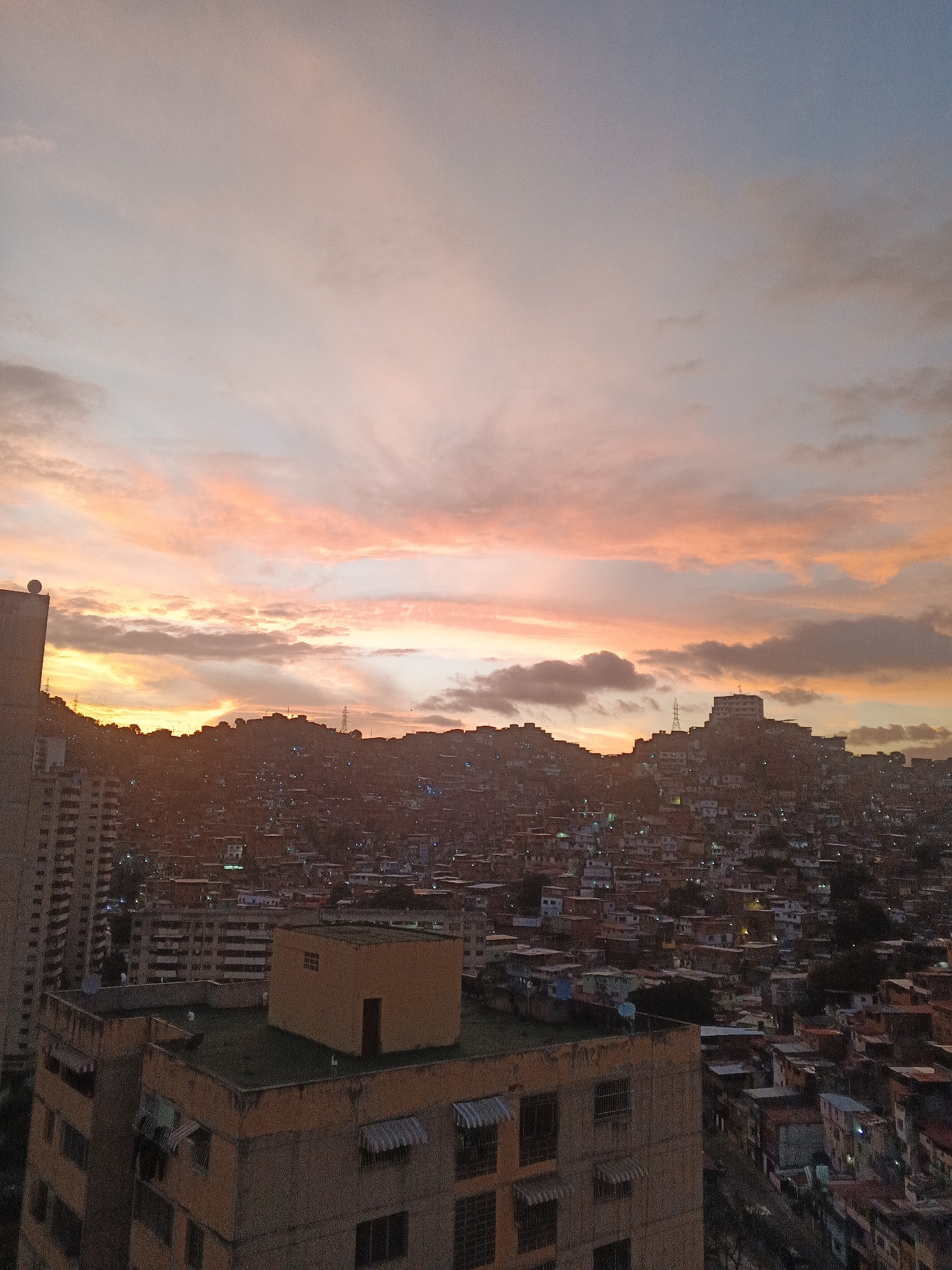 The image shows a cityscape during a sunset or sunrise, likely in Caracas, Venezuela.
**Environment:**
* Sky is filled with pink, orange, and light blue hues indicative of sunrise or sunset.
* Hilly terrain in the background is densely populated with housing.
**Subjects & Objects:**
* Foreground features several buildings with flat roofs and windows.
* A large building, potentially an apartment block, is on the left side of the frame.
* The city in the middle ground is composed of tightly packed, varied-sized buildings and dwellings.
**People & Actions:**
* No people are clearly visible in the shot.
* Lights are visible in some buildings, suggesting it is either twilight or early morning.
**Interactions:**
* The scene implies urban life within a densely populated area.
* No direct interactions are depicted, but the environment indicates human activity.