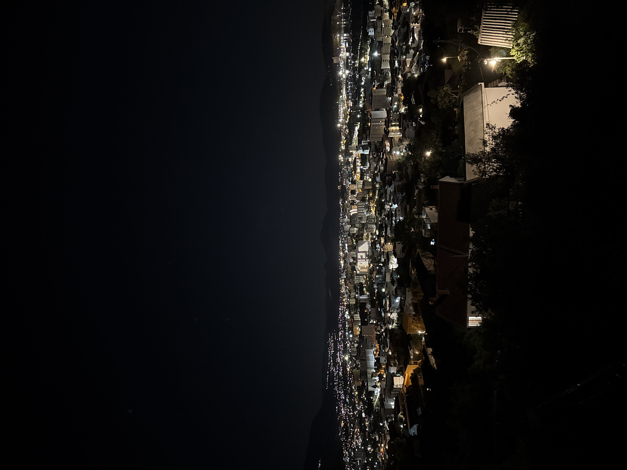 This is a nighttime view overlooking Šušanj, Montenegro. The scene is divided between the dark expanse of the Adriatic Sea and the illuminated cityscape stretching along the coastline. The city lights reveal buildings of varying sizes and densities, concentrated along the coast and fading into the dark hills. In the foreground, a few structures are visible, including what appear to be residential buildings with distinct rooflines. The perspective suggests the photograph was taken from an elevated vantage point, likely a nearby hillside or mountain. There are no visible people or specific activities.