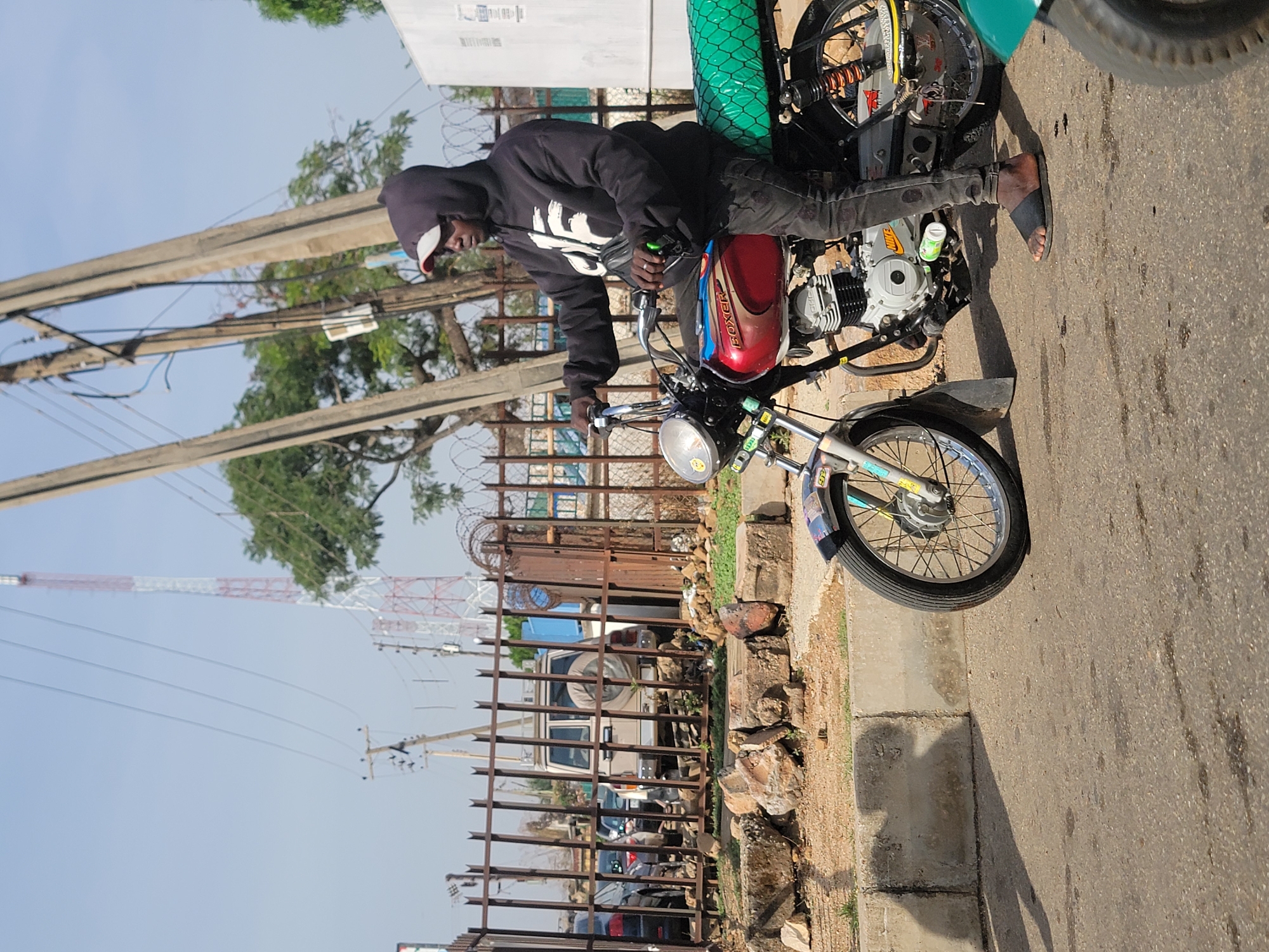 The image shows a man sitting on a motorcycle in an urban environment in Jos, Nigeria. The man wears a black hoodie and dark pants, with one foot on the ground in a sandal. The motorcycle has a red and maroon fuel tank, a green-covered cargo rack, and chrome accents. In the background, there is a rusted metal fence, parked vehicles, utility poles with wires, trees, and a communications tower in the distance. The setting appears to be a street or roadside with a curb and sidewalk.