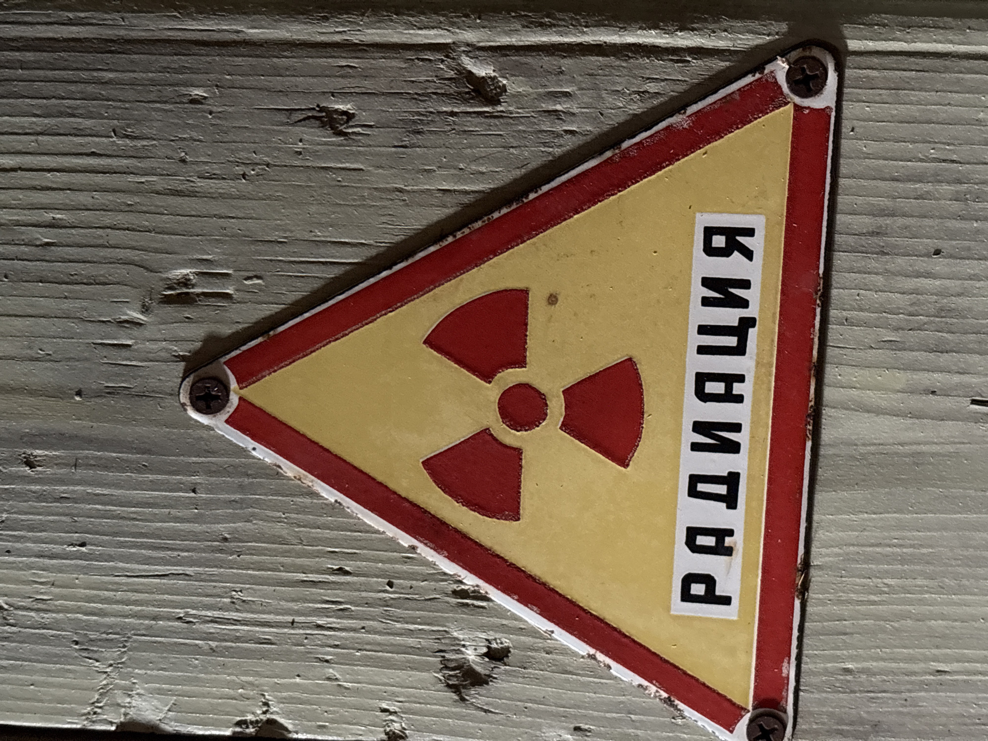 The image shows a yellow triangle with a red border and a red trefoil symbol in the center, indicating radiation. Below the symbol, the word "РАДИАЦИЯ" (radiation in Russian) is written in black letters on a white rectangle. The sign is affixed to a weathered, light-colored wooden surface with screws. The texture of the wood suggests an outdoor or unmaintained environment. The image is a close-up of the sign and a portion of the wooden surface it is attached to. No specific location in the city of Yaiva, Russia, is identifiable from the image itself.