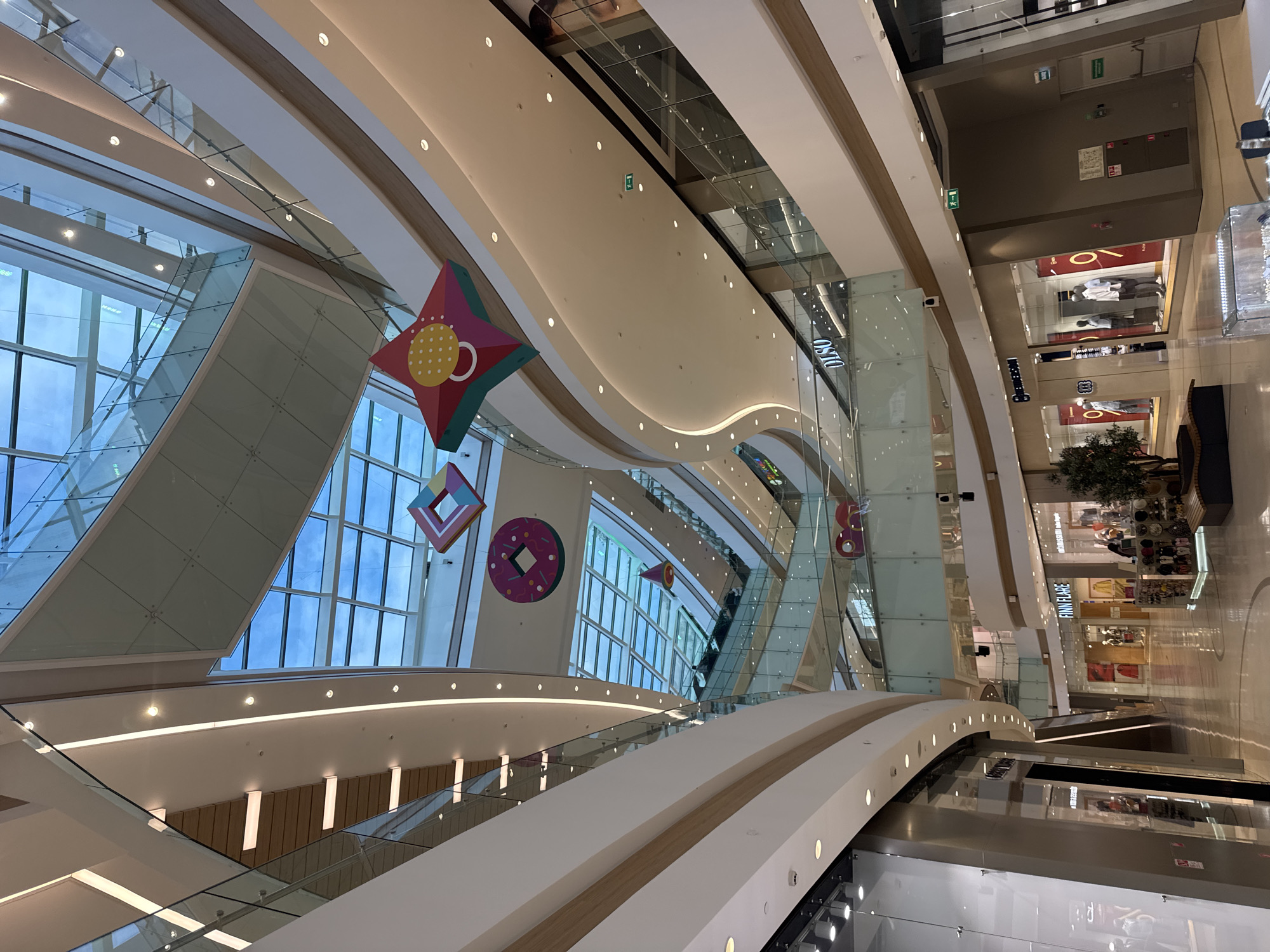 The image shows the interior of a modern, multi-level shopping mall. The architecture is characterized by sleek, curved lines, extensive use of glass and white surfaces, and a large, transparent ceiling that allows natural light to flood the space. Decorative geometric shapes, resembling a star, a donut, and a diamond, are suspended from the ceiling. Escalators and walkways connect the different floors, with glass railings providing views into the various retail levels. Shops with illuminated storefronts are visible, displaying mannequins and merchandise. Signs for "FINN FLARE" and "DLSO" are present. The flooring appears to be polished, reflecting the lighting and architecture. The overall atmosphere is bright, airy, and contemporary. The location is specified as Moscow, Russia.