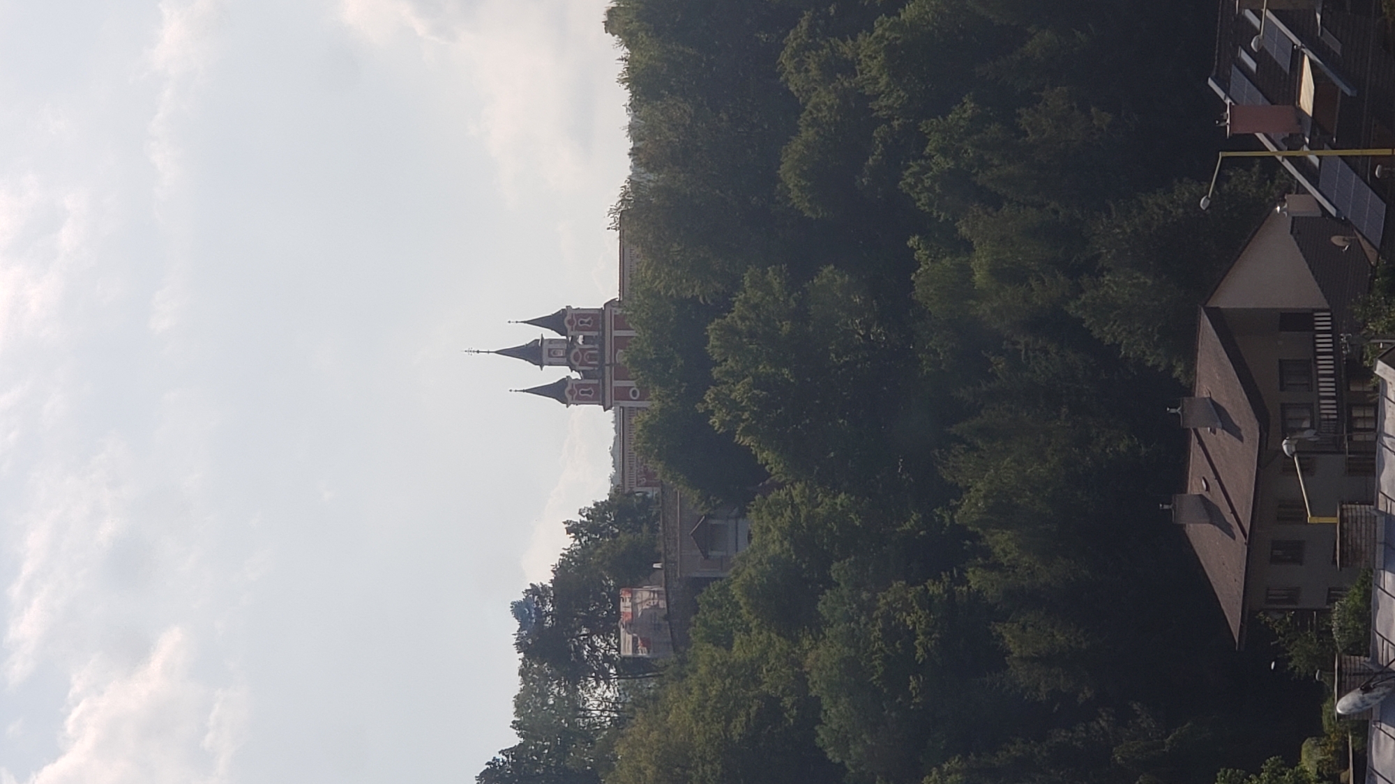 The image shows a view from a distance of a red building with multiple spires, possibly a church or monastery, situated on a hill covered in trees. In the foreground, there are several houses with pitched roofs, some equipped with solar panels and satellite dishes. The sky is partly cloudy. The building with spires is identified as the Catholic Church of the Holy Spirit in Prešov, Slovakia, a historical landmark in the city.