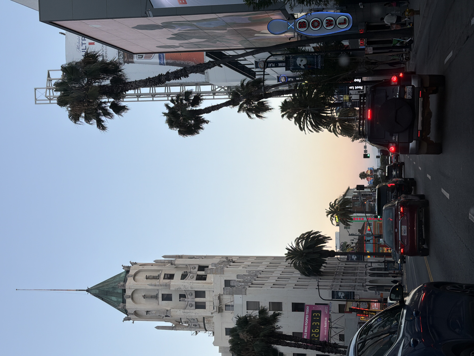 The image depicts a street scene in Hollywood, Los Angeles, USA. The sky is a pale gradient of blue and orange, indicating dusk or dawn. Dominating the left side of the frame is the distinctive Art Deco architecture of the Eastern Columbia Building, with its green pyramidal roof and clock face. Palm trees line the street, interspersed with buildings showcasing signage for various businesses and attractions, including "PHANTOM THE OPERA" and "PANTAGES". Several cars are driving on the street, and a few pedestrians are visible on the sidewalks. The overall atmosphere suggests a busy urban environment in a well-known entertainment district.