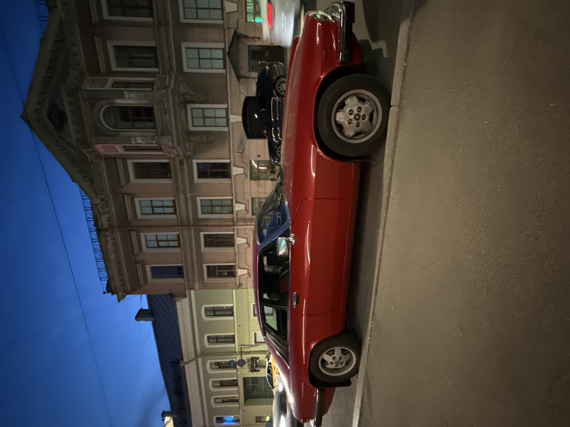 A red vintage Jaguar coupe is parked on a street in Saint Petersburg, Russia, during twilight. The car is viewed from the side, showcasing its classic design. In the background, ornate buildings with classical architecture line the street. The sky above is a deep blue, indicating the late evening hour. Other cars are visible on the street, some in motion, creating streaks of light due to the long exposure. The scene captures a moment of urban life in a historic European city.
