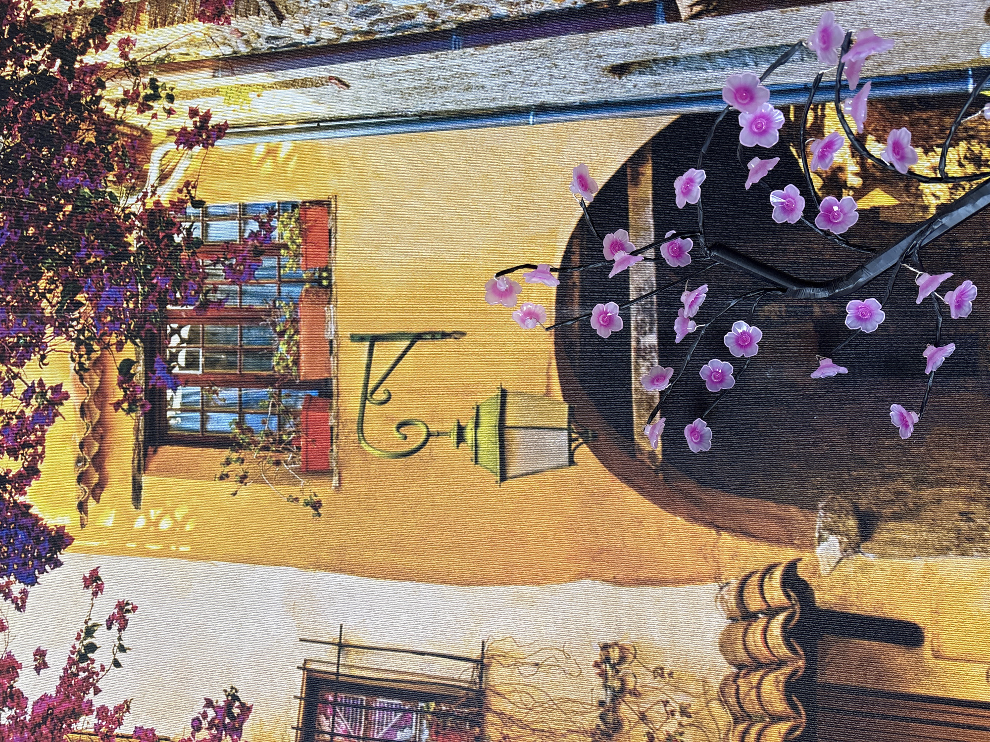 This is a detailed, close-up, fabric tapestry image depicting a scene of a European-style building facade with architectural elements and floral accents. The building features a vibrant yellow-ochre wall, a dark arched doorway, and a window with a wooden frame and red flower boxes. Above the window, a traditional green street lamp is mounted on an ornate metal bracket. The scene is enhanced by a large branch with numerous translucent pink artificial flowers in the foreground, creating a whimsical and decorative effect. The upper portion of the tapestry shows a cascade of purple flowers, likely bougainvillea, suggesting a Mediterranean or Southern European setting. The texture of the tapestry is visible, characterized by interwoven threads. The image does not contain any people or specific identifiable landmarks that would place it in Georgiyevsk, Russia.