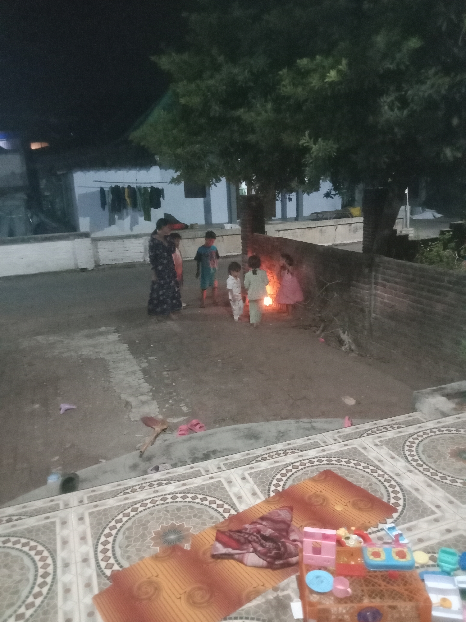 In Kedungsari, Indonesia, at dusk, several individuals, including children and at least one adult woman, are present in an outdoor courtyard. The ground is made of packed dirt and concrete, with decorative tiled flooring visible in the foreground. Several buildings, including one with laundry hanging outside, form the backdrop. A low brick wall runs through the scene. A warm, orange glow emanates from a light source near a group of children, suggesting a lamp or lantern. Scattered on the ground are various objects, including pink sandals, a small purple object resembling a toy, and a toy kitchen set in the foreground. The overall scene depicts a casual, communal gathering in a residential outdoor space.