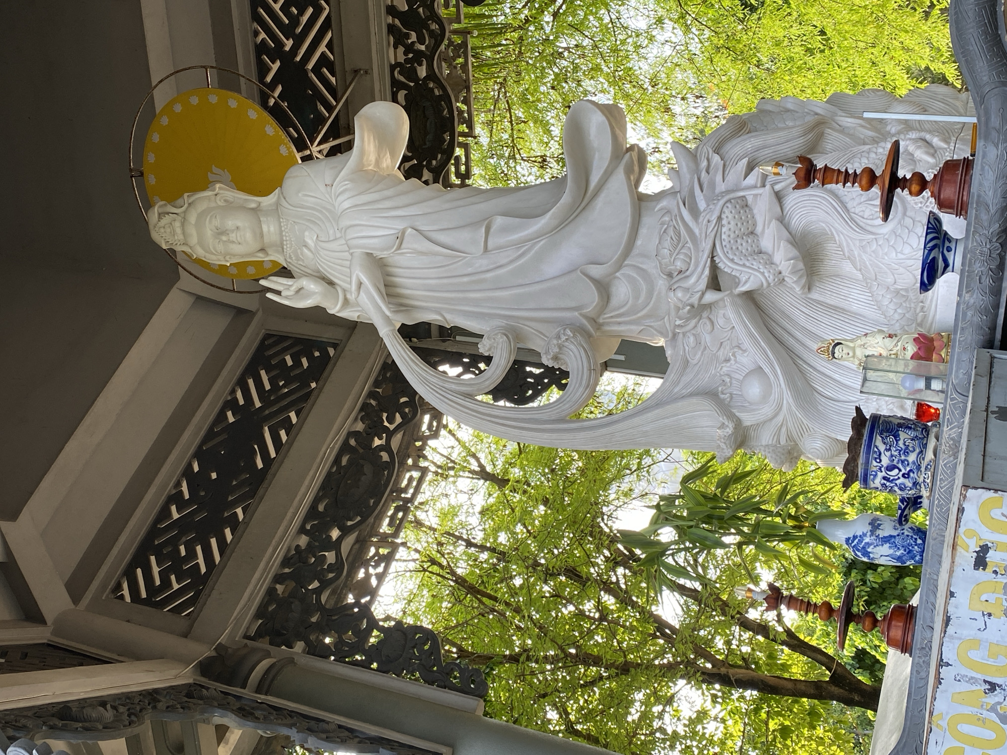 This image captures a large, white marble statue of Guan Yin, the goddess of mercy, positioned under a decorative pavilion in Tay Nha Trang Ward, Vietnam. The statue stands on a base adorned with a dragon. Guan Yin is depicted with flowing robes and is holding an object in her right hand, with her left hand raised in a blessing gesture. A yellow halo with lotus petal designs is visible behind her head.
In front of the statue, on a stone altar, are several offerings. These include a wooden candle holder with a lit candle, a blue and white porcelain vase, a blue and white ceramic pot with an incense burner, a small figurine of a Buddha, and a red glass object. The altar also has a weathered sign with yellow lettering.
The background is filled with lush green trees, suggesting an outdoor setting. The sunlight filters through the leaves, creating a bright and airy atmosphere. The pavilion structure itself is ornately carved, with dark wood elements visible. The overall scene conveys a sense of religious devotion and tranquility.