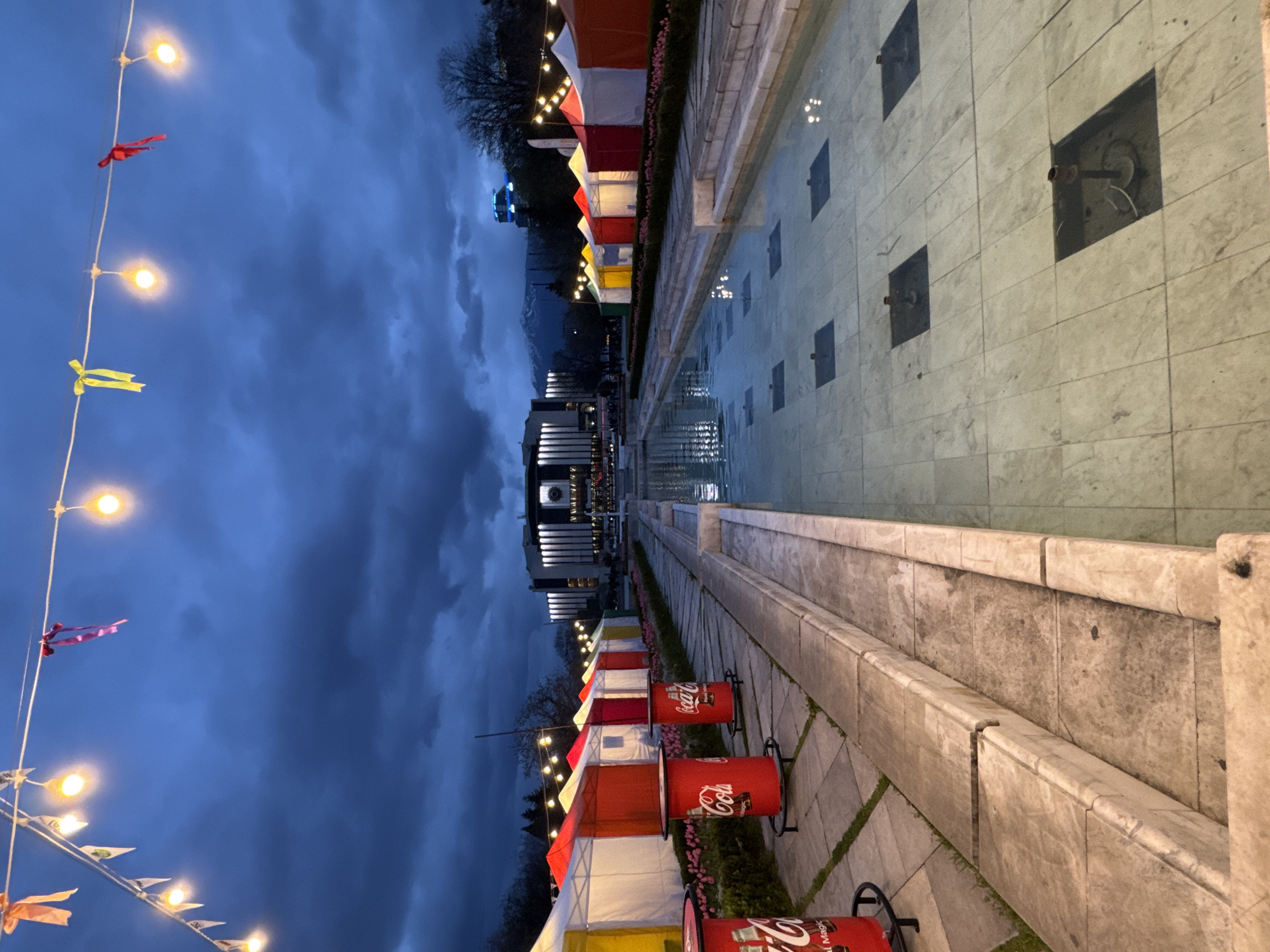 The image shows a view of a city square in Sofia, Bulgaria, likely taken at dusk or early evening. A long, narrow reflecting pool runs through the center of the scene. On either side of the pool are paved walkways. Along one side are small, brightly colored tents or stalls, some with Coca-Cola branded displays. Strings of lights with colorful pennants are strung above the walkway. In the distance, a large building can be seen, possibly a public building or cultural center. The sky is cloudy and dark. The scene appears to be set up for a festival or market. There are no people visible in the image.