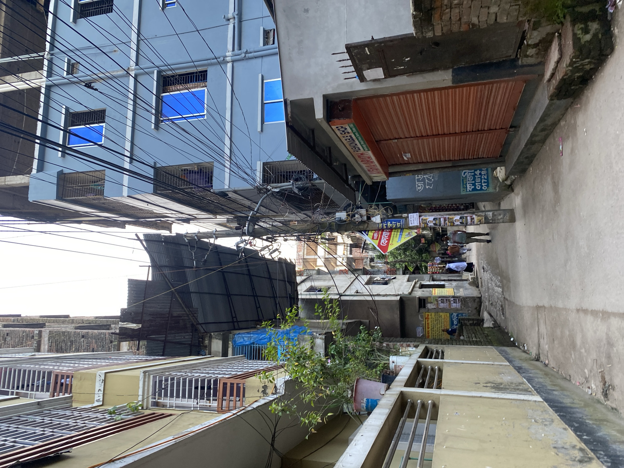 The image depicts a narrow street in Dhaka, Bangladesh, bordered by multi-story buildings on both sides. The buildings display a mix of architectural styles and states of repair, with some appearing newer and painted in pastel colors, while others show signs of age and disrepair, with exposed brickwork and makeshift additions. Wires are strung haphazardly across the street, a common sight in the city.
On the left, a pale yellow building with white trim features balconies with metal railings and some plants in pots on a ledge. Adjacent to it is a darker building with metal grating. Further down the street, a building is covered with a blue tarp, and a corrugated metal roof extends over a section of the alley.
On the right, a light blue building has several windows, some with blue-tinted glass and metal security bars. Below this building, a shop with a rolled-down orange shutter is visible, with a sign above it reading "AVSHA ARATH FASHIONABLE SHOP". Another shop to the right has a dark metal door.
Several people are present in the alley. Two individuals are walking away from the camera towards the background. One person in the mid-ground appears to be wearing a blue shirt and is standing near a wall covered in posters. Another person in the background is wearing a white hijab.
The street itself is paved but appears somewhat uneven and dirty, with some debris scattered on the ground. The overall impression is of a densely populated urban environment with a vibrant, if somewhat chaotic, atmosphere.