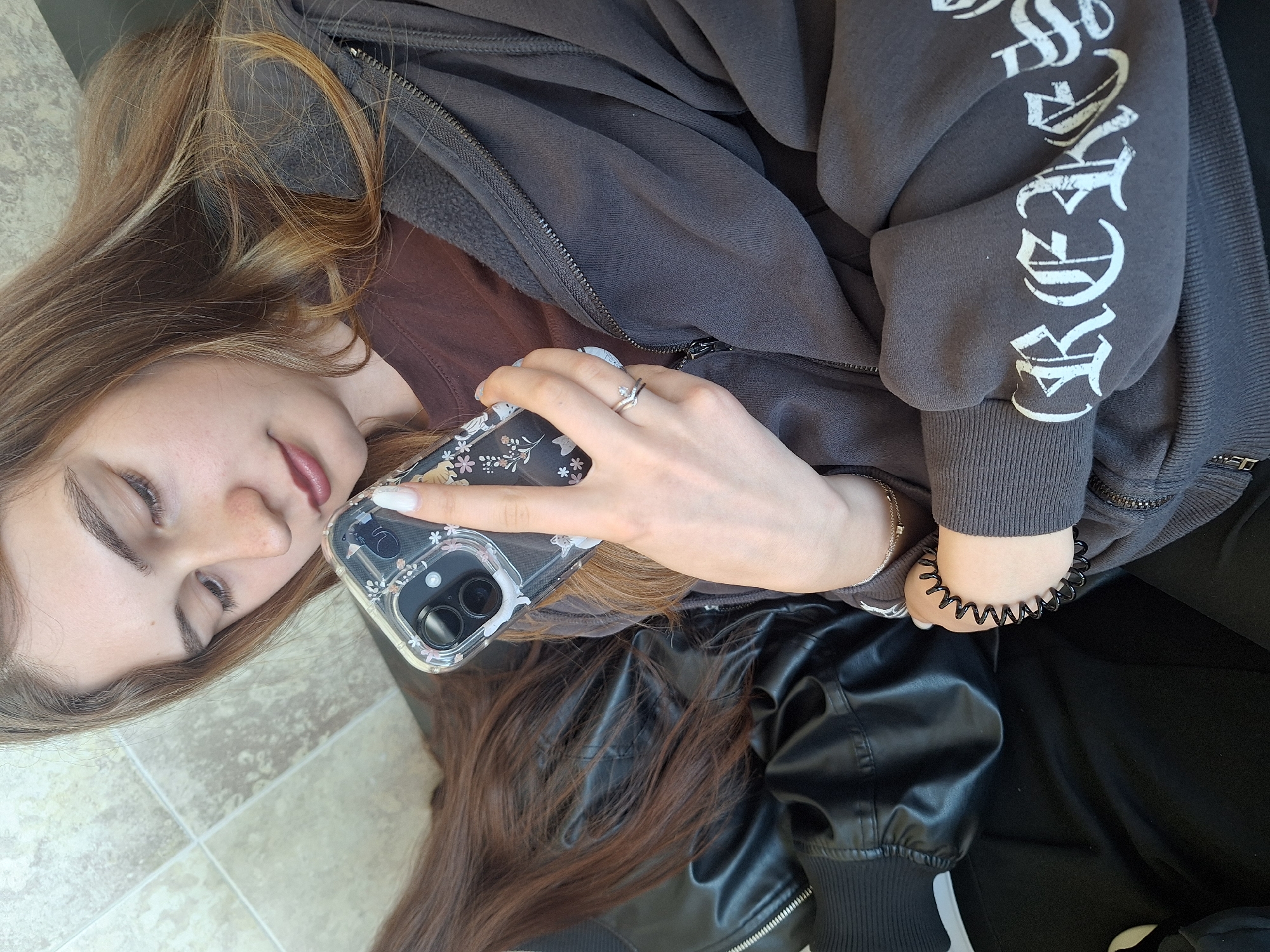A young woman with long brown hair lies on a tiled surface. She is holding a smartphone with a clear case decorated with flowers and cats. She is wearing a dark gray zip-up hoodie with white lettering on the sleeve and a brown t-shirt underneath. She has a silver ring on her finger. She also has a black spiral hair tie on her wrist. Part of a black leather jacket with white trim is visible at the bottom of the frame. The location is unknown.