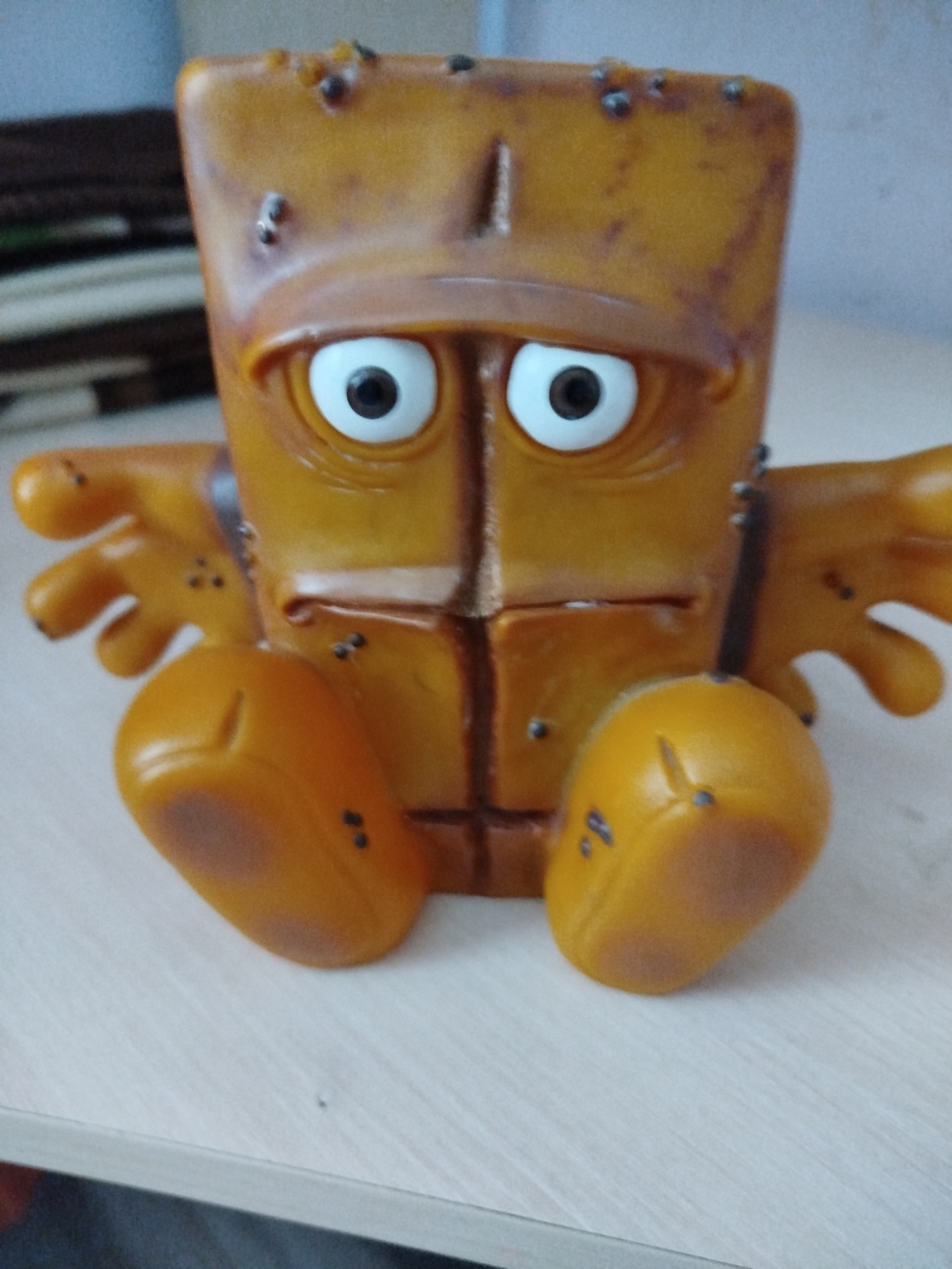 A close-up, eye-level shot shows a worn, orange plastic toy character sitting on a light-colored wooden surface. The toy is square-shaped with a protruding brim over its eyes, resembling a baseball cap. It has two large, white, circular eyes with black pupils, and a rectangular mouth. Small, dark dots are scattered across its head and body, giving it a somewhat textured or dirty appearance. The toy has small, detached arms extending outwards from its sides, and two rounded lower limbs that appear to be its legs. The background is blurred, with hints of darker colors and a light blueish-grey hue at the top left. The scene does not contain any people or indicate a specific location within Крывасельскі сельскі Савет, Belarus.