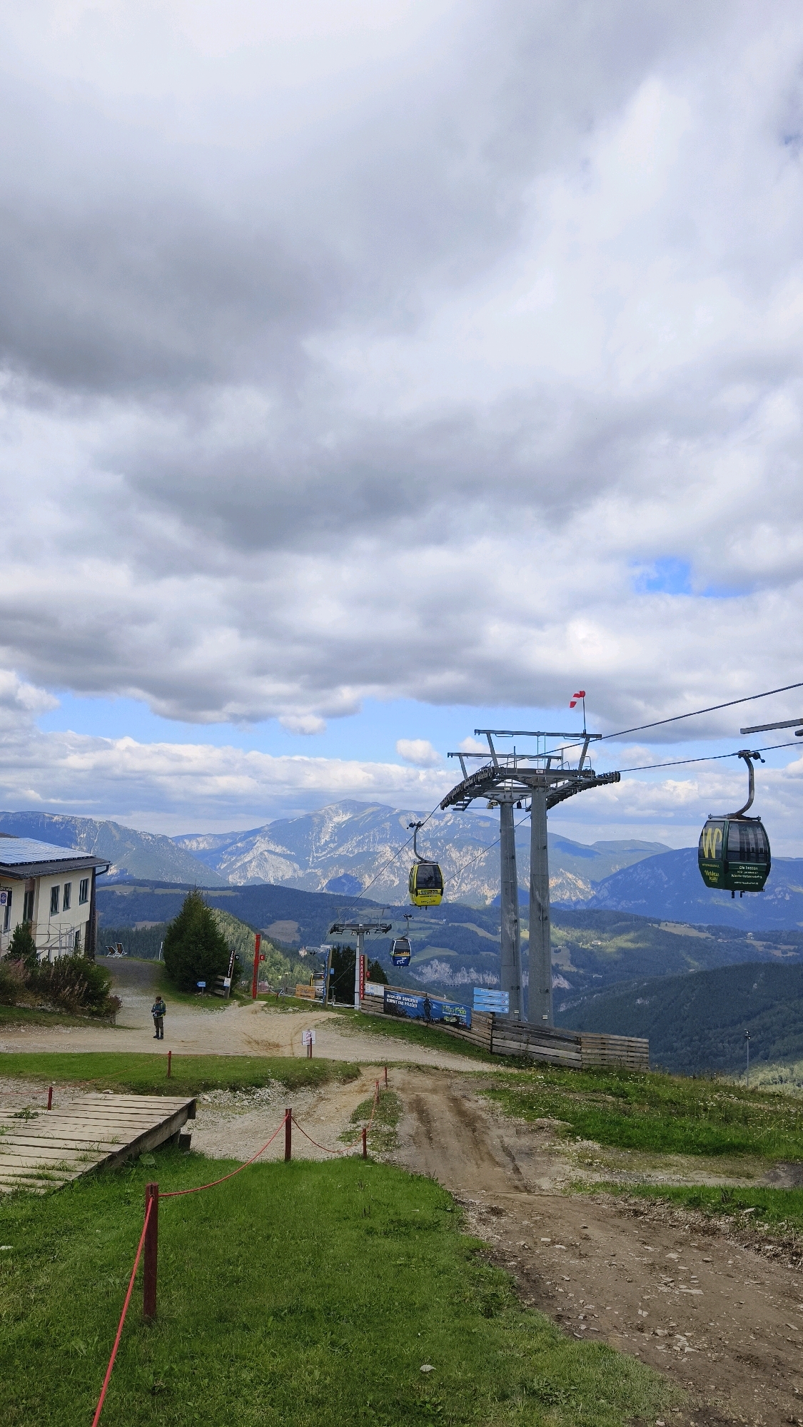The image depicts a mountainous landscape in Semmering, Austria, characterized by a cable car system and a building. In the foreground, a dirt path curves to the right, bordered by grassy areas and a wooden walkway on the left. Red ropes delineate sections of the path and grassy areas. A single person, wearing dark clothing, stands on the path near a building. The building, located on the left side of the frame, has a light-colored facade and solar panels on its roof.
In the midground, the cable car towers rise, supporting the cables of the gondola lift. Two gondolas are visible: one yellow and one dark green, each with branding. The yellow gondola has "Wiener Linien" written on it, while the green gondola has "WIENER WALZER" and a logo. A small Austrian flag is attached to the top of one of the cable car towers.
The background showcases a vast mountain range with layered peaks, some of which are covered in trees and others appear rocky. The sky is filled with a mix of white and dark grey clouds, with patches of blue sky visible. The overall scene suggests an outdoor recreational area, likely a ski resort or a scenic mountain viewpoint.