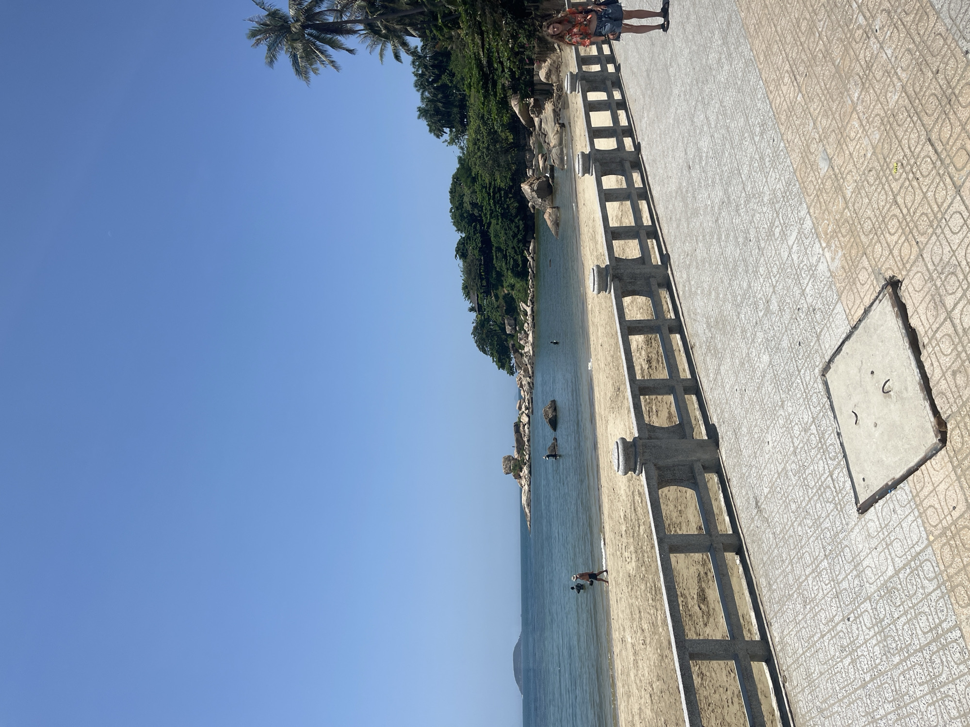 A bright, sunny day at a beach in Nha Trang, Vietnam. The image captures a panoramic view of the coastline, with the azure sea meeting the sandy shore. In the foreground, a paved walkway runs parallel to the beach, separated by a stone balustrade. A young woman in a colorful top and denim shorts stands on the walkway, facing the sea. Further down the beach, a man in a straw hat walks along the water's edge, while other figures can be seen wading in the shallows. Rocky outcrops and lush greenery dot the coastline, with palm trees swaying gently in the breeze. The clear blue sky stretches across the horizon, completing the idyllic tropical scene.