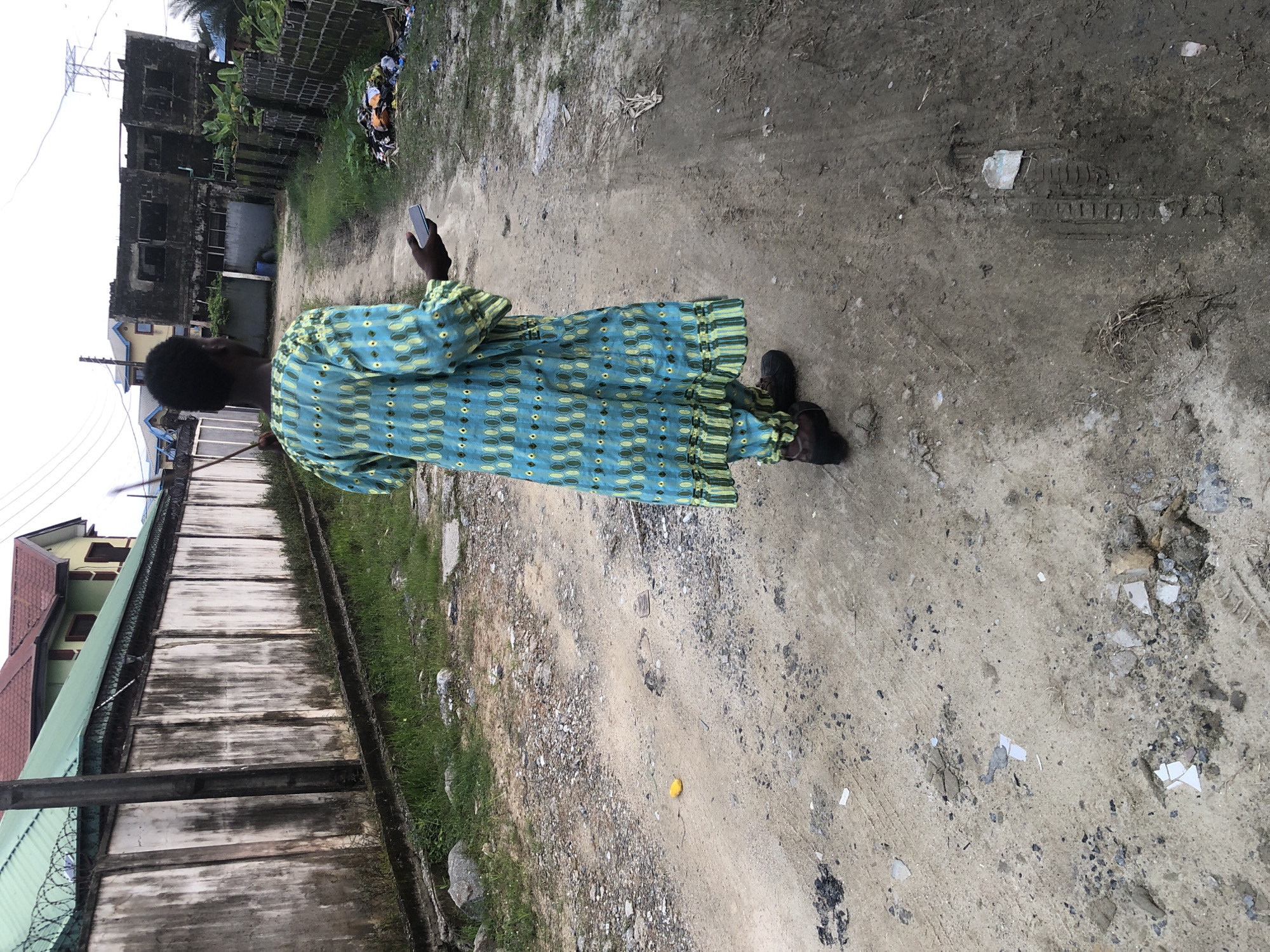 A lone individual stands on a dirt path, facing away from the camera and holding a mobile phone. They are wearing a blue and green patterned traditional outfit with matching pants and a long-sleeved tunic, along with dark shoes. The path appears to be in a rural or semi-urban area, with overgrown grass and debris lining the sides. In the background, there is a weathered concrete wall with barbed wire on top, a building with a red roof, and more distant structures that appear to be abandoned or under construction. The sky is overcast, suggesting a cloudy day. The environment is somewhat unkempt, with scattered trash visible on the right side of the path. The scene seems to depict a quiet moment in a developing or neglected part of Ado, Nigeria.