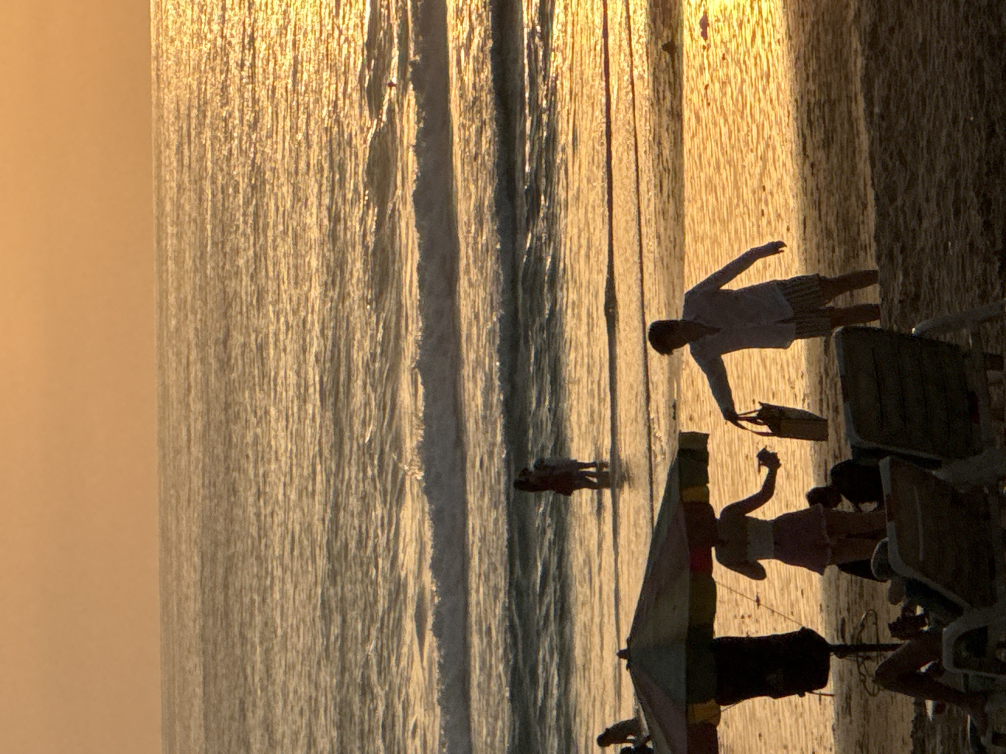 The image depicts a beach scene at sunset in Karon, Thailand. The sky is a warm orange, casting a golden glow over the ocean and sand. Several people are visible, silhouetted against the bright water. In the foreground, two beach chairs are occupied by individuals lounging. To the left, an umbrella shades a portion of the sand, with a dark bag hanging from its pole. A woman in a light-colored top and a pink skirt stands near the water's edge, holding something in her hand. A man in a white shirt and striped shorts is walking towards her, carrying a tan bag. Further out in the water, two pairs of people are standing, appearing to interact. Gentle waves lap the shore, reflecting the sunset's light. The overall atmosphere is tranquil and serene.