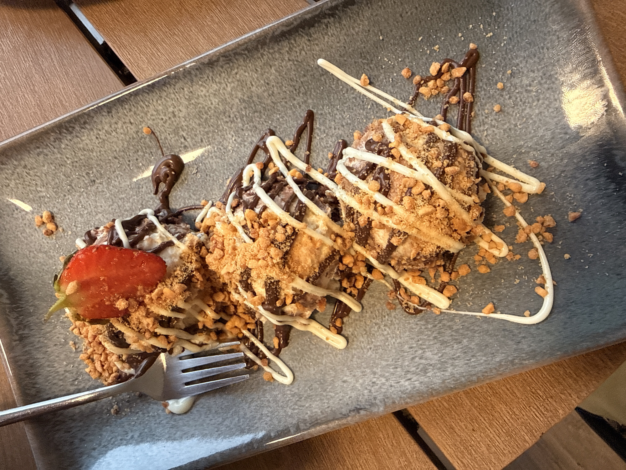 A top-down, medium shot shows a dessert on a grey, rectangular plate. The dessert is composed of what appears to be pieces of waffle or pastry, drizzled with dark chocolate and white icing, and sprinkled with crushed nuts or cookies. A single strawberry slice is placed on top of the dessert. A fork is resting on the plate, partially over the dessert. The plate is positioned on a wooden surface, possibly a table. The lighting suggests an indoor setting. The image does not contain any identifiable landmarks or specific details to determine a location within Lovech, Bulgaria.