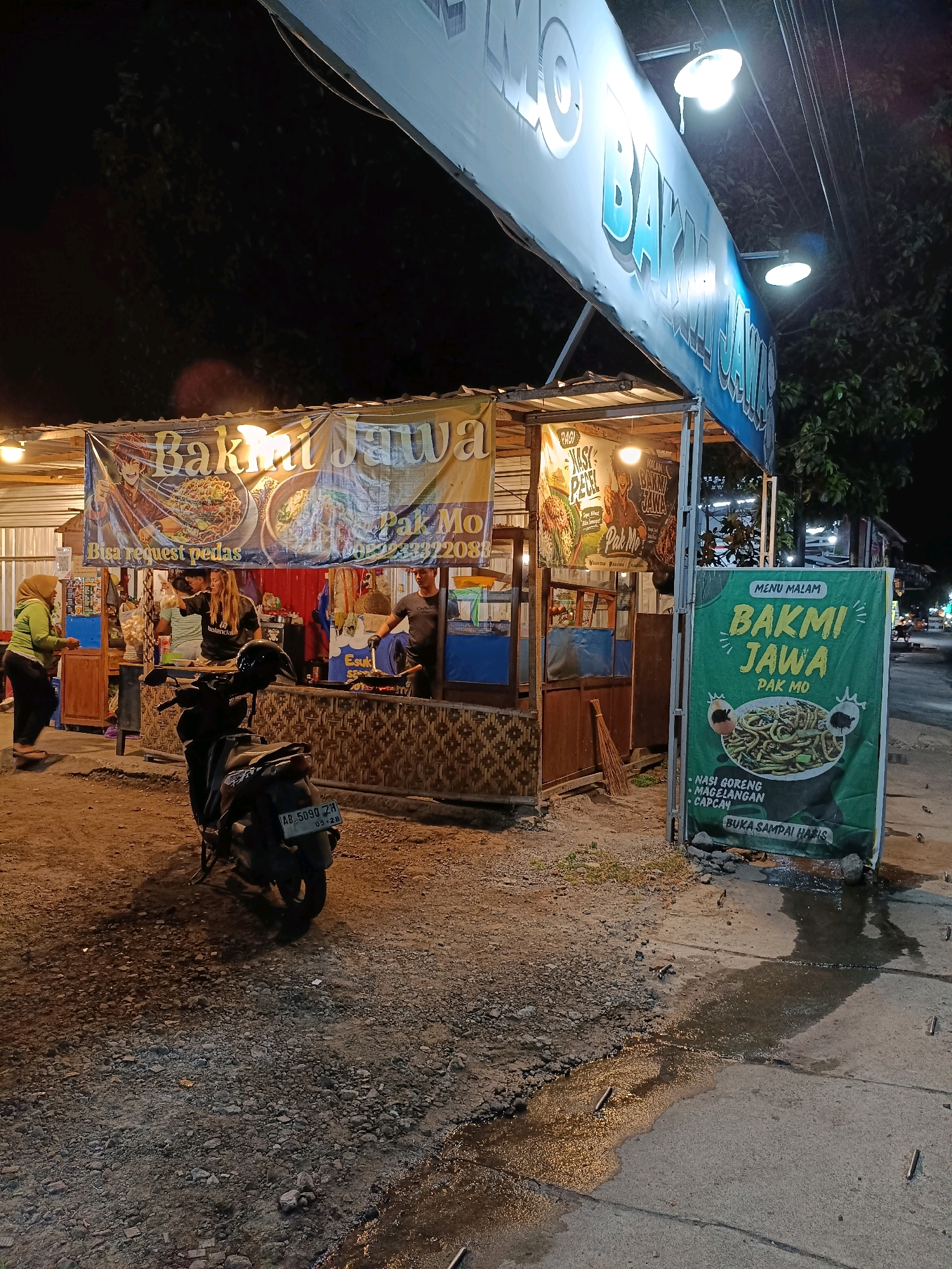 At night in Maguwoharjo, Indonesia, a "Bakmi Jawa Pak Mo" food stall is illuminated, featuring a prominent banner with the text "MENU MALAM BAKMI JAWA PAK MO" and an image of noodles. To the left, a smaller banner advertises "PAGI NASI PECEL PAK MO". Three staff members are visible behind the counter: a woman in a black t-shirt, a man in a grey t-shirt, and another individual whose face is obscured. A woman in a green hijab and black pants stands near the entrance of the stall. A dark-colored motorcycle with license plate AB 5090 ZH is parked in front of the stall. The ground is a mix of dirt, gravel, and some standing water reflecting the stall's lights. The overall scene depicts a typical street food vendor operating after dark.