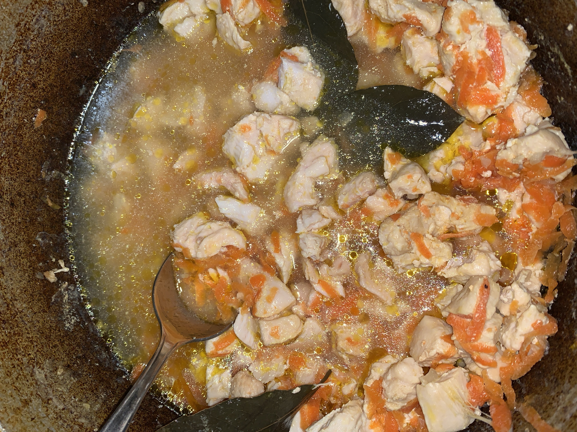 A high-angle, close-up shot depicts a pot of cooked food, likely a stew or braise. The food consists of pale, cubed pieces of what appears to be cooked chicken or tofu, mixed with thinly sliced orange vegetables, possibly carrots. The food is submerged in a light-colored broth with visible oil droplets. Two bay leaves are partially visible, resting on top of the food. A stainless steel spoon sits in the pot, partially submerged. The pot itself appears to be made of a dark, possibly cast iron material, and shows signs of wear and use. The location is Unknown, Unknown.